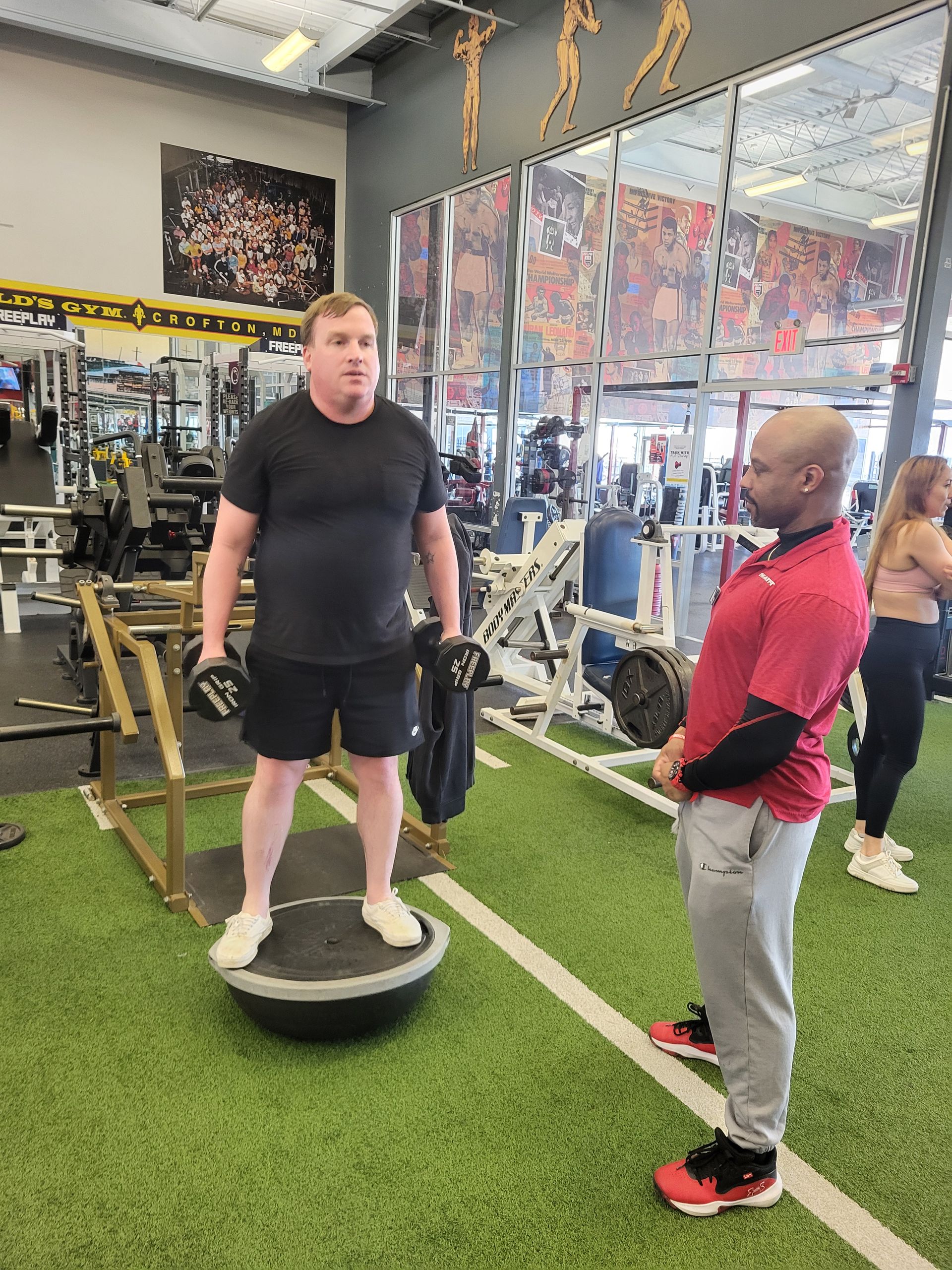 Man on balance trainer holding dumbbells, being instructed by another man in a gym.