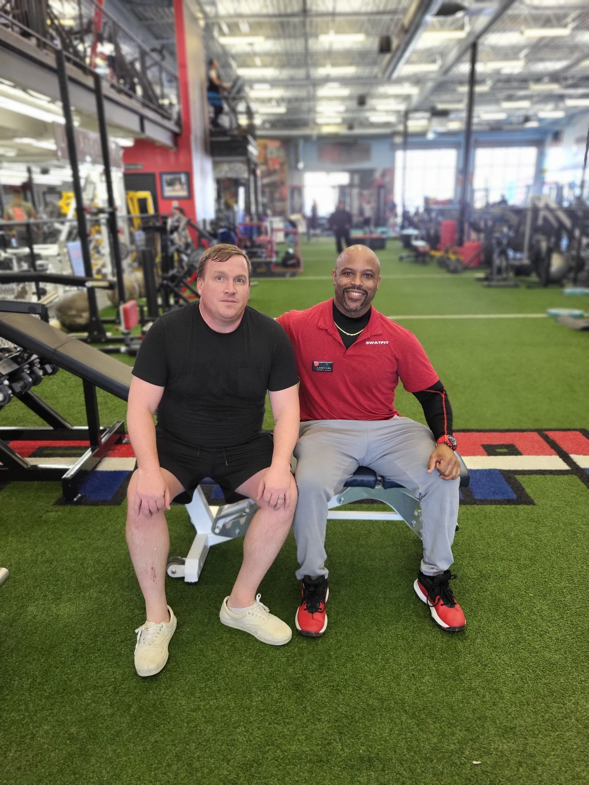 Two men seated on a bench in a gym. One wears black, the other a red shirt. They both smile.