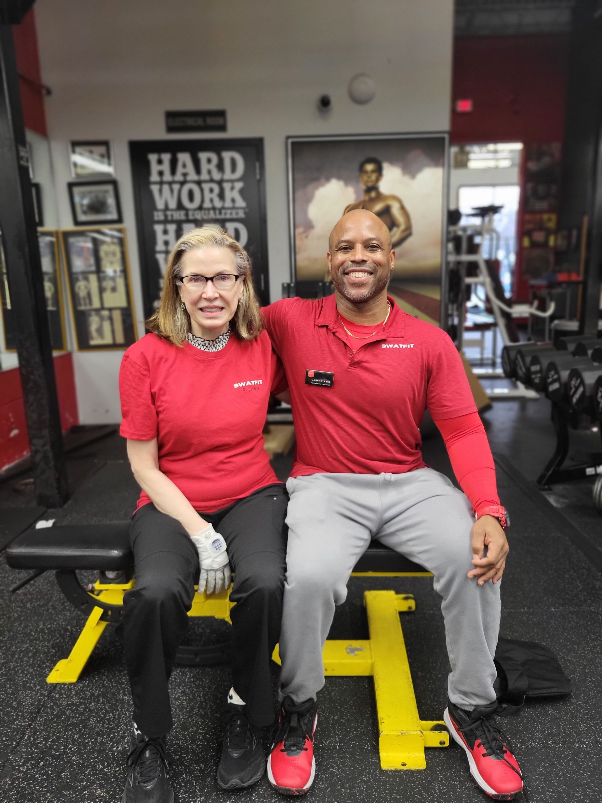 Two people in red shirts pose on a weight bench in a gym.  