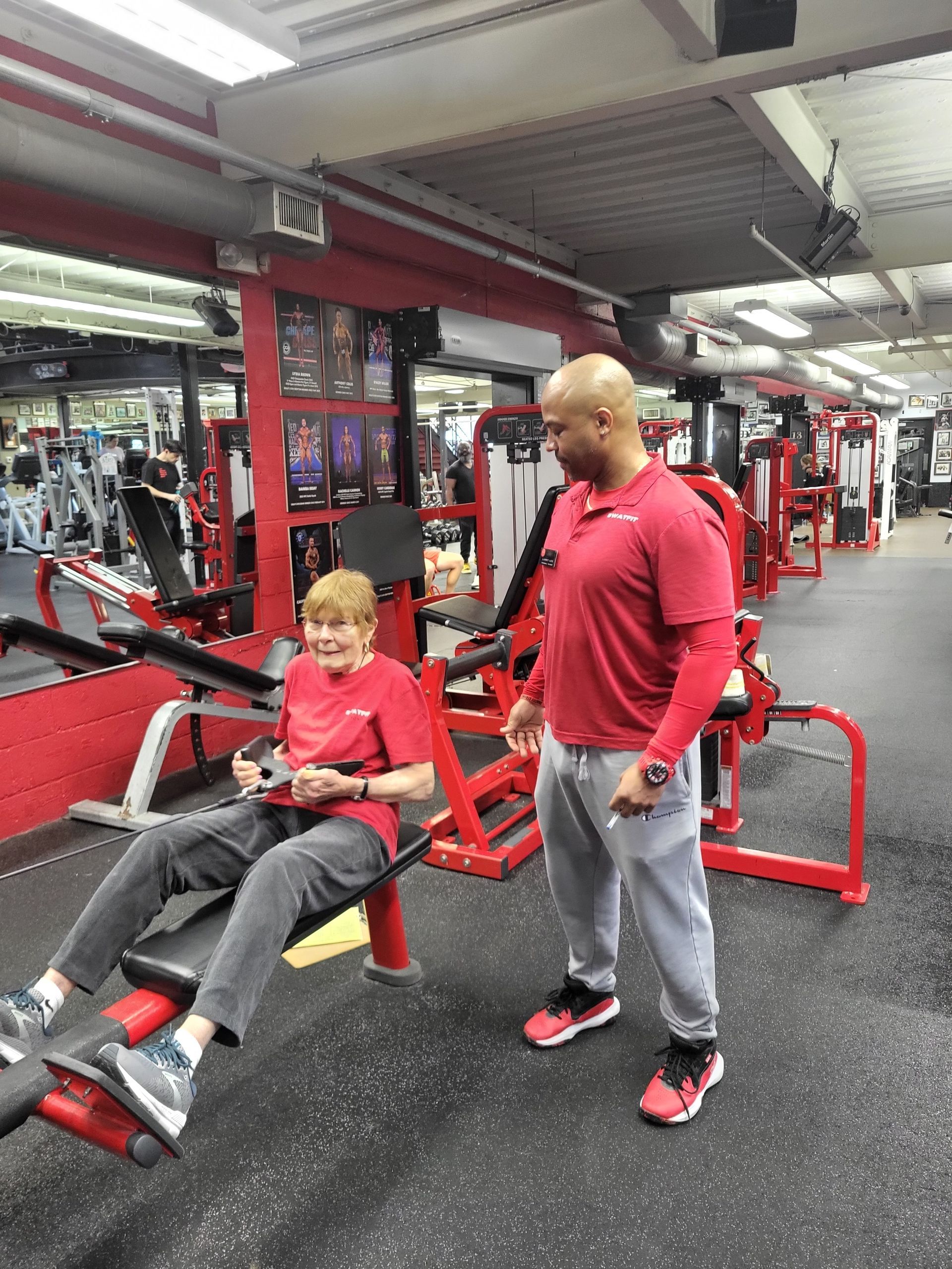 A person using gym equipment with an instructor. Both wear red shirts. Gym setting with red machines.