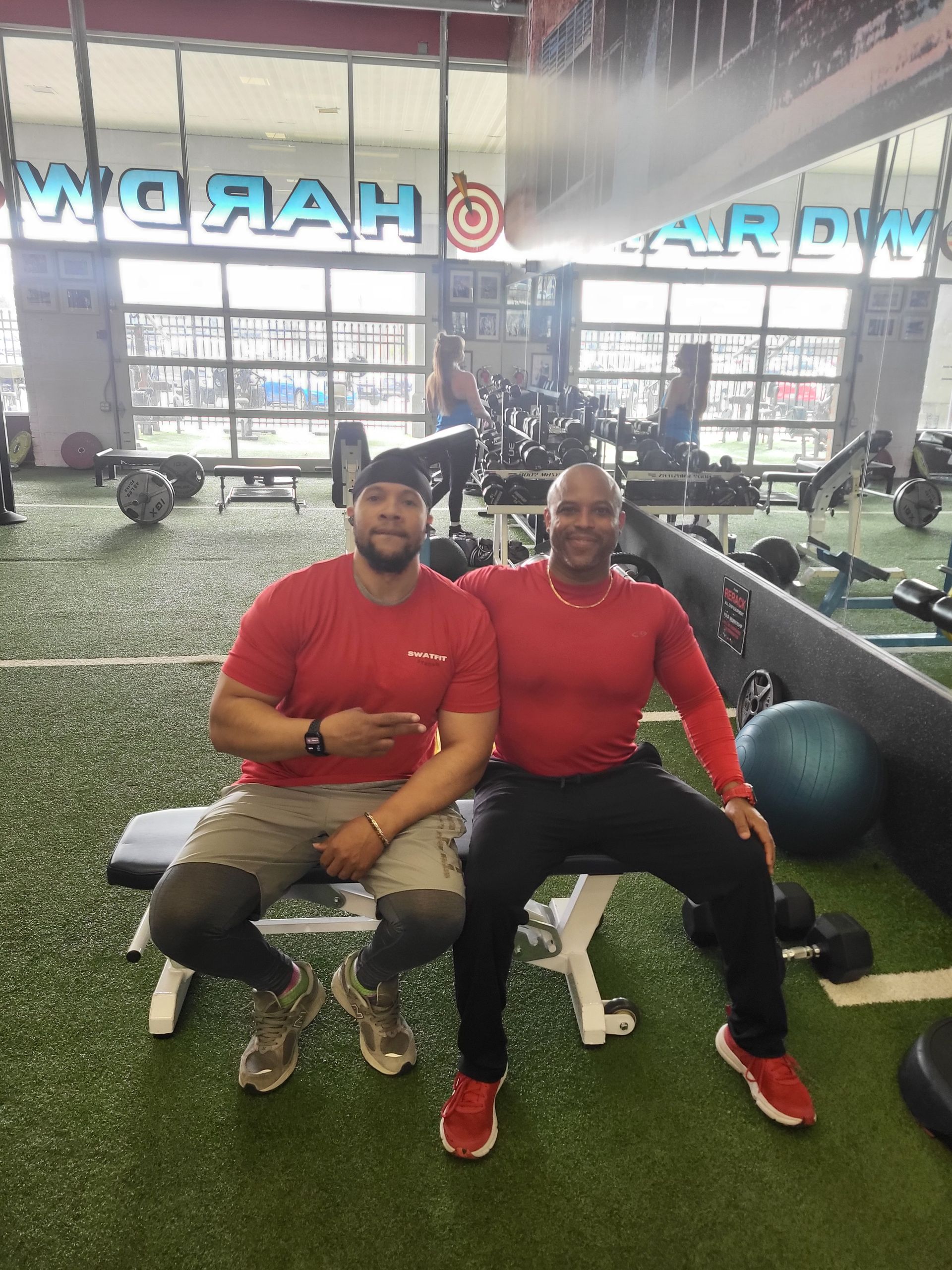 Two men in red shirts sit on a gym bench. One points; the other smiles. They are in a gym.
