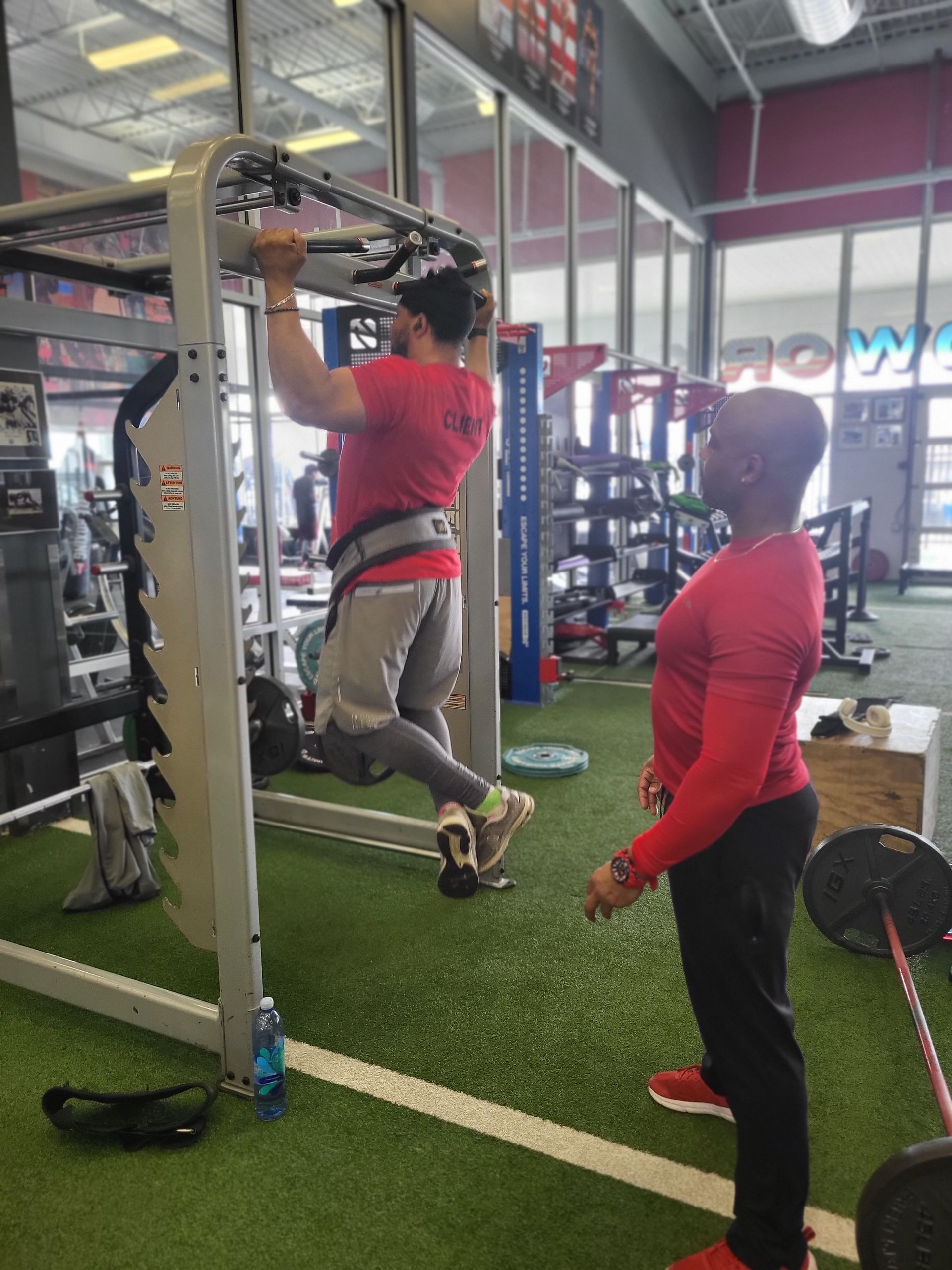 Man doing pull-up with weight belt at gym, with another man observing. Green turf, red and gray clothing.