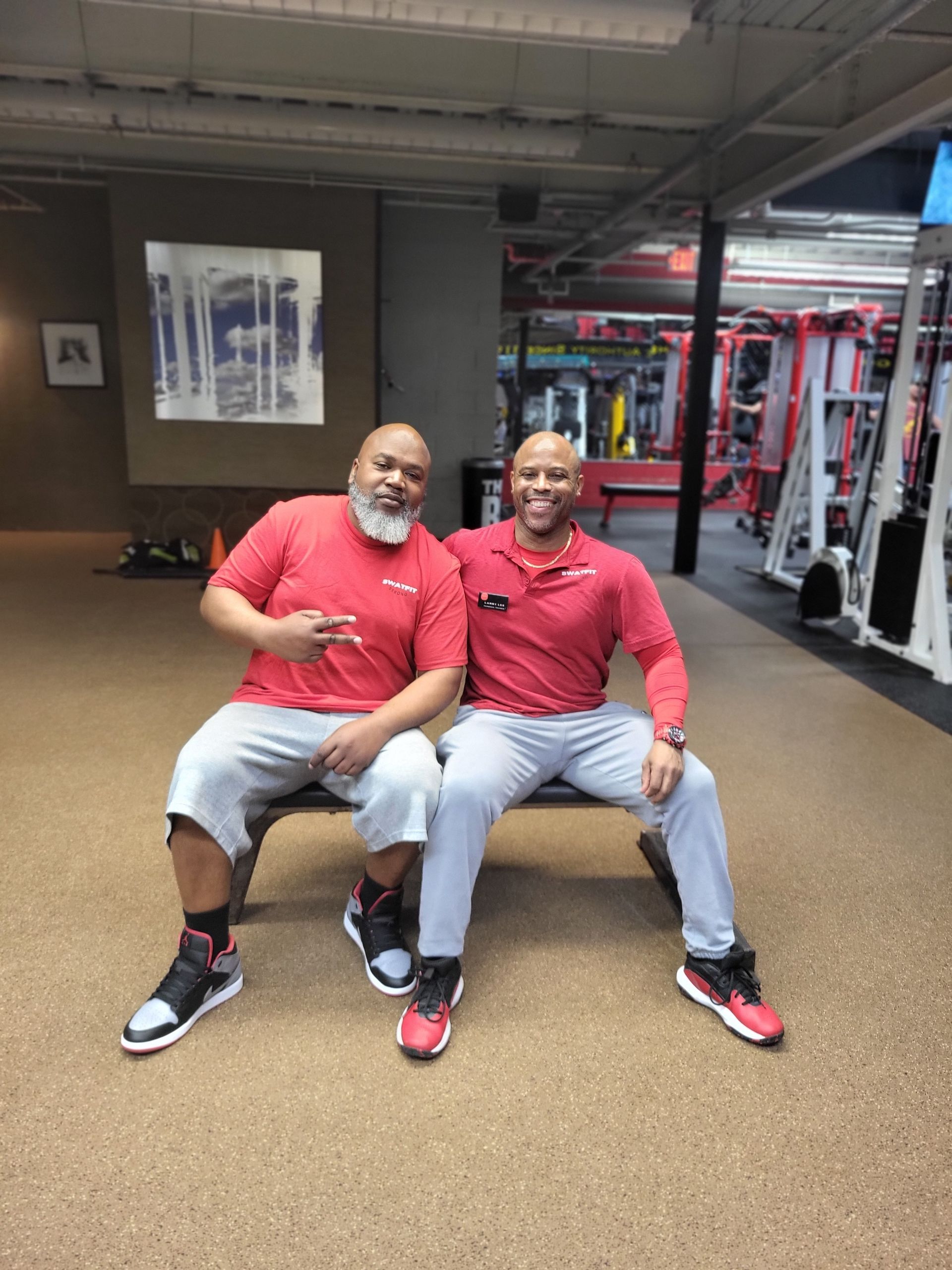 Two men in red shirts and gray pants, seated on a bench in a gym. One man is gesturing a peace sign.