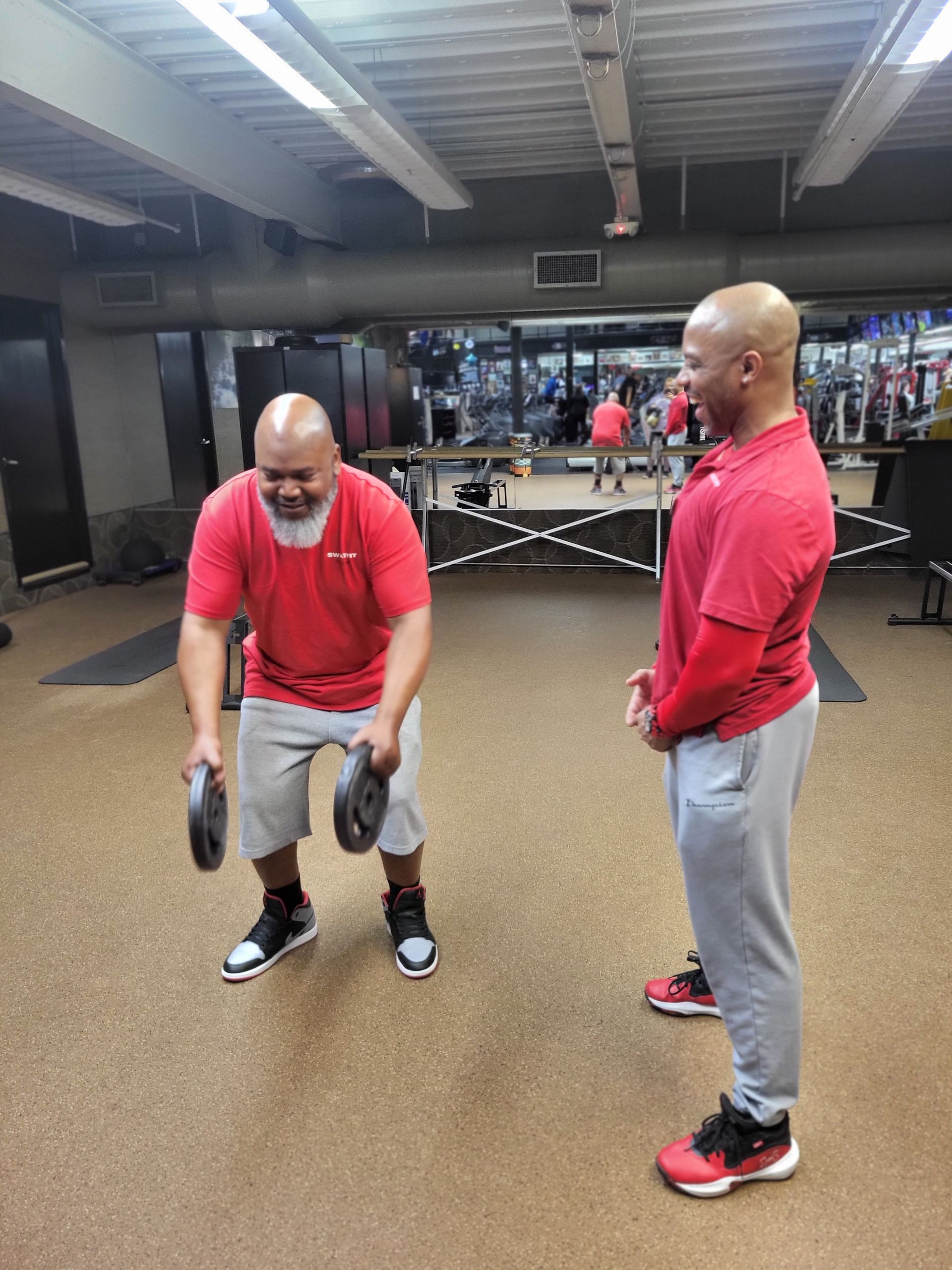 Man exercising with weights, coach observes. They're in a gym. Both wear red shirts and athletic shoes.