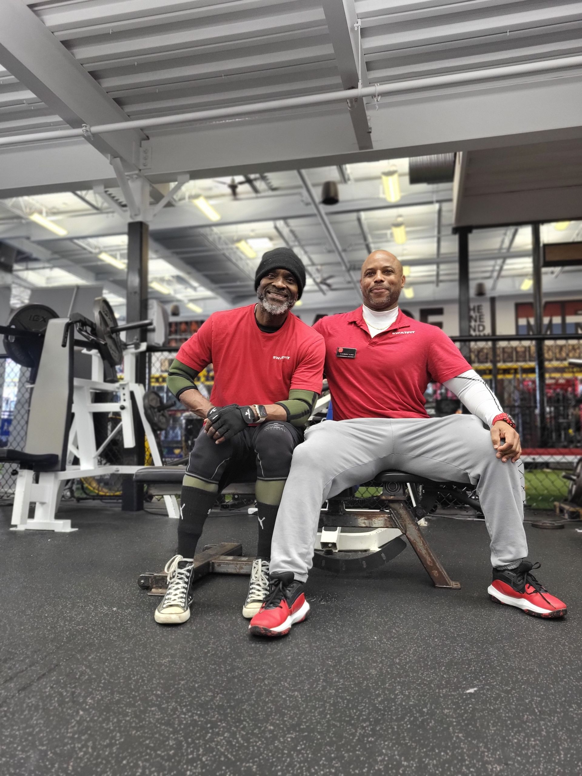 Two men in red shirts sit on a weight bench in a gym. One has a beard, the other is smiling.