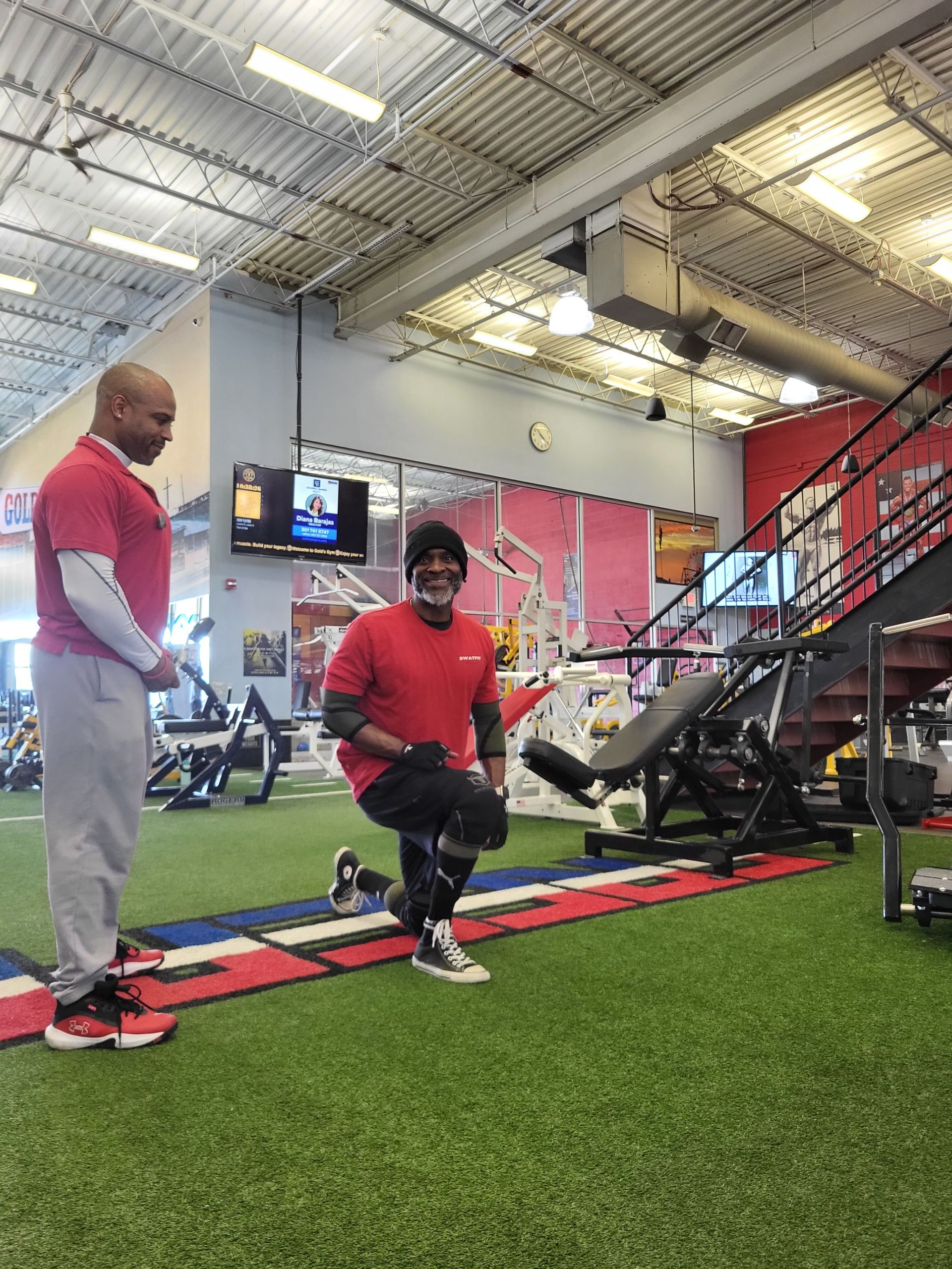Person performing a lunge exercise in a gym with a trainer nearby. Artificial turf, red and gray accents.