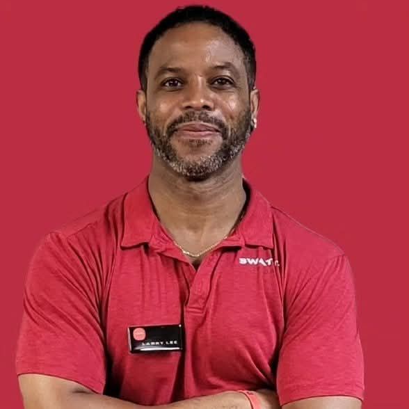 Man wearing red polo shirt, nametag, arms crossed, smiling, red background.