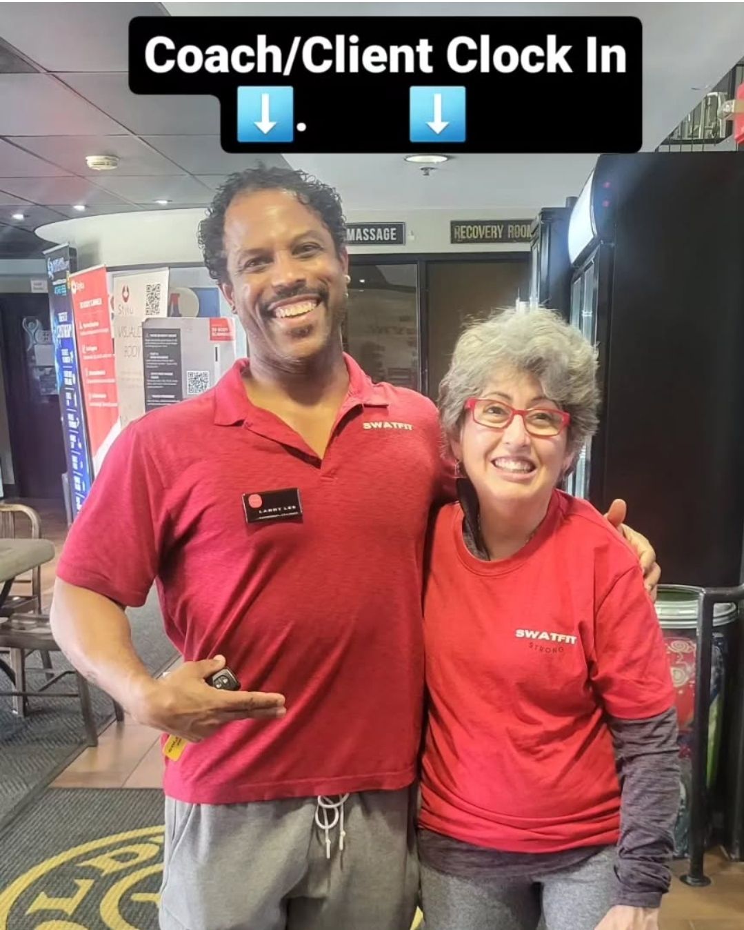 Two people in red shirts at a gym, smiling. One points down. 