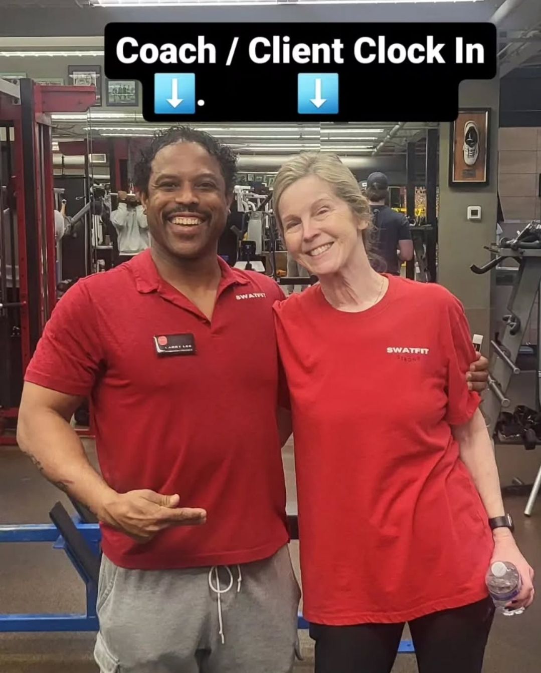 A coach and client in red shirts pose in a gym. Coach points, both smile.