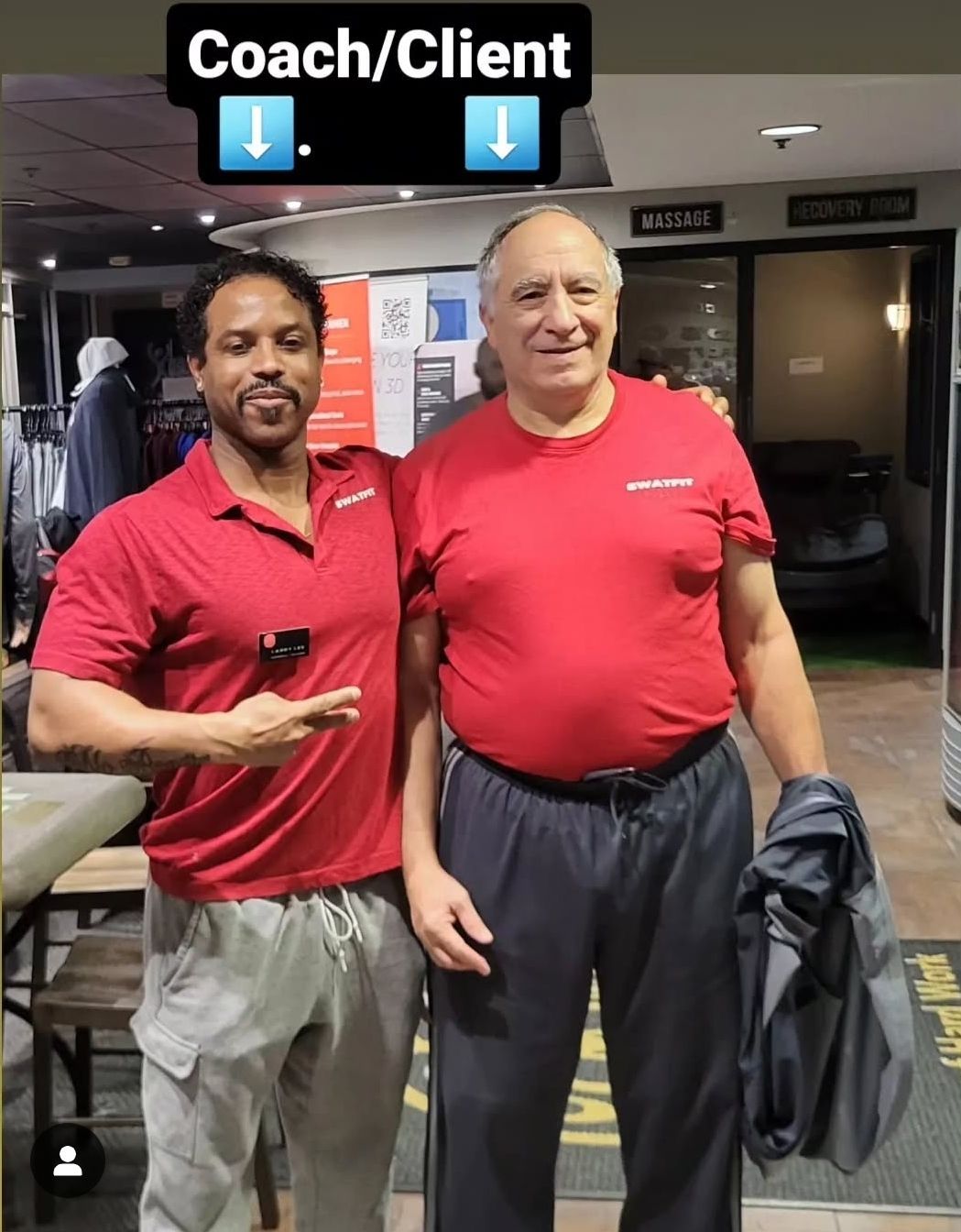 Coach and client standing together. Coach pointing. Both wearing red shirts. Gym setting.