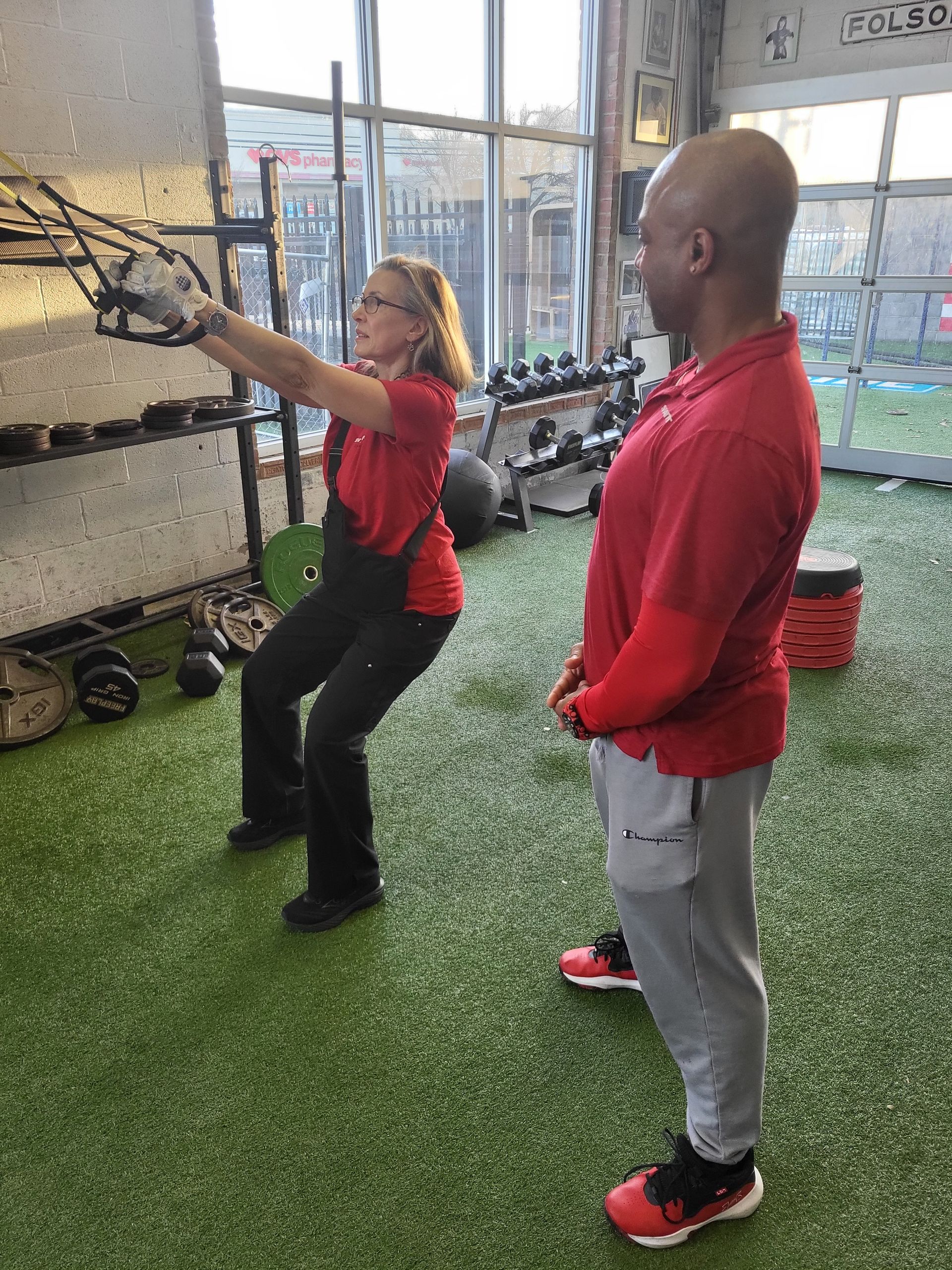 Woman in a gym doing a squat with trainer observing. Both wear red shirts, gym setting with equipment visible.