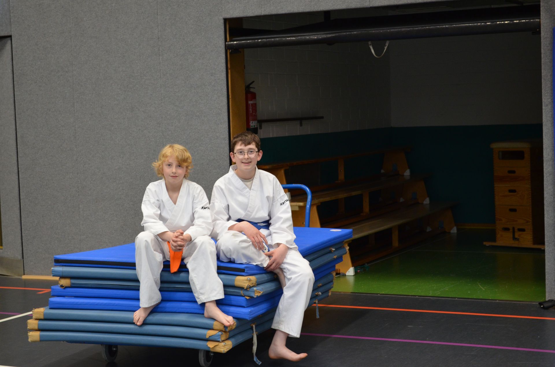 Zwei kleine Jungen sitzen in einer Turnhalle auf einem Stapel blauer Matten - Karate-Jugendfreizeit.