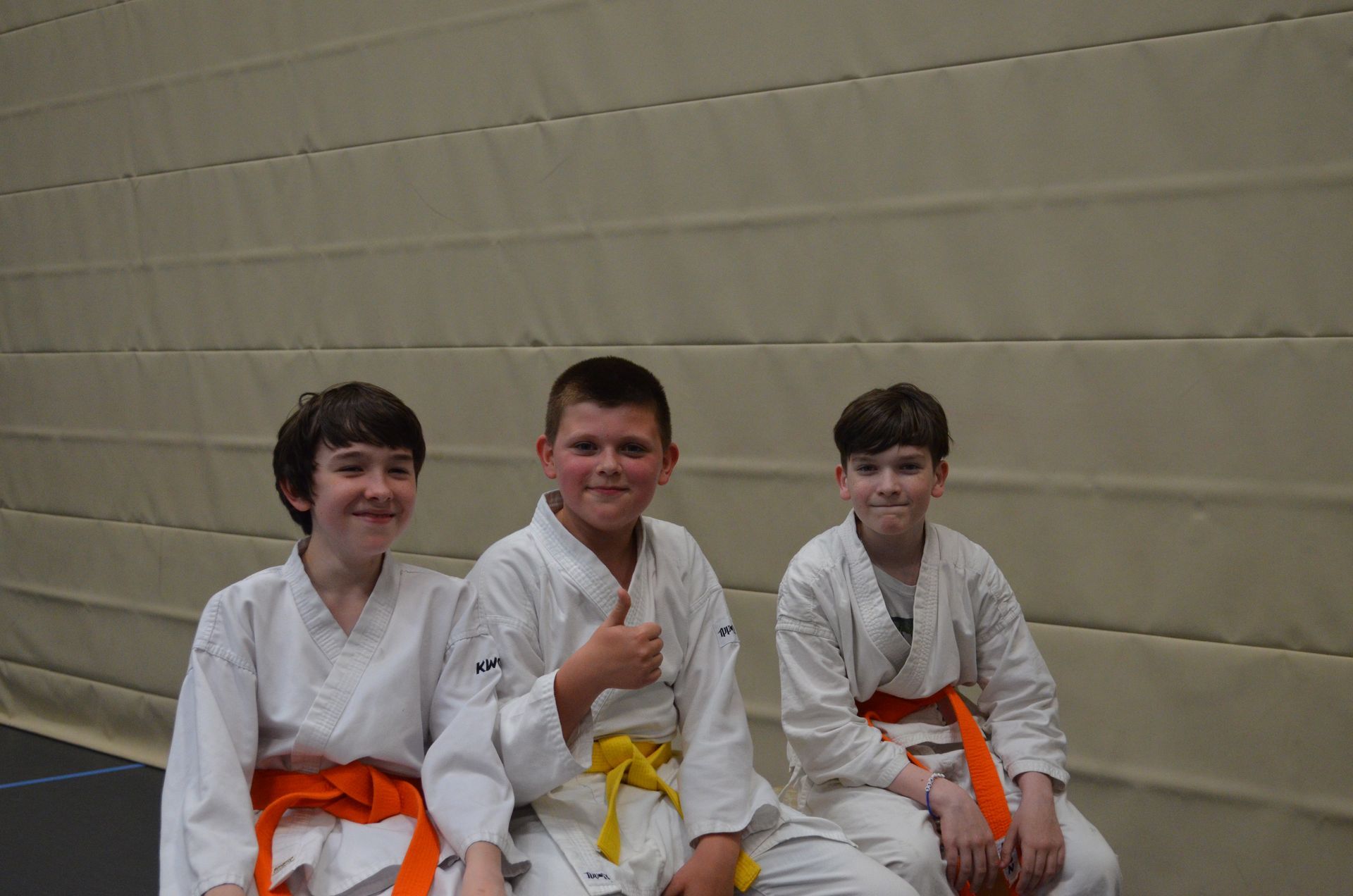 Drei junge Jungen in Karateuniformen sitzen nebeneinander und strecken den Daumen nach oben - Karate-Jugendfreizeit.