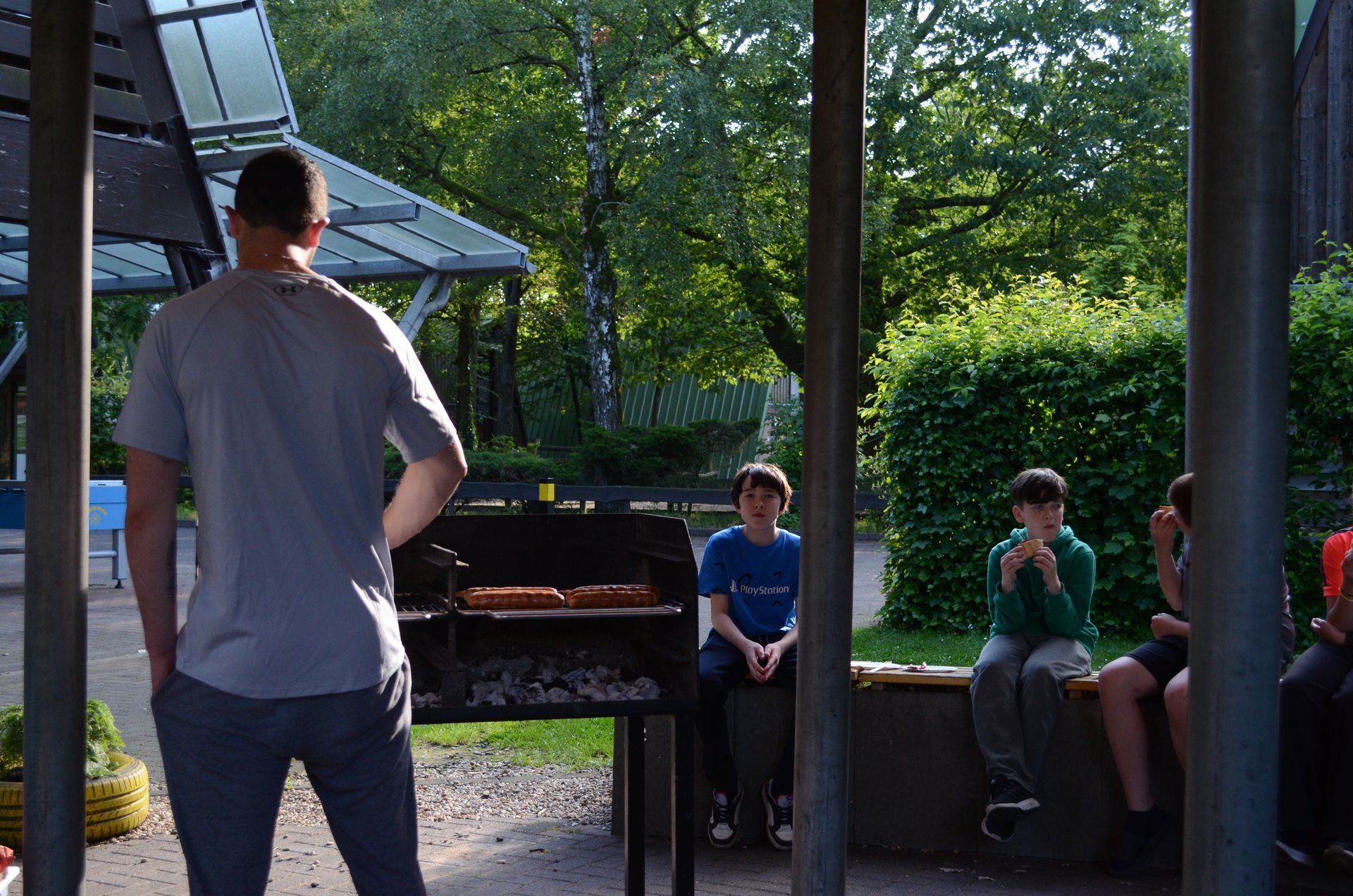 Ein Mann steht vor einem Grill und eine Gruppe Leute sitzt darum herum - Karate-Jugendfreizeit.