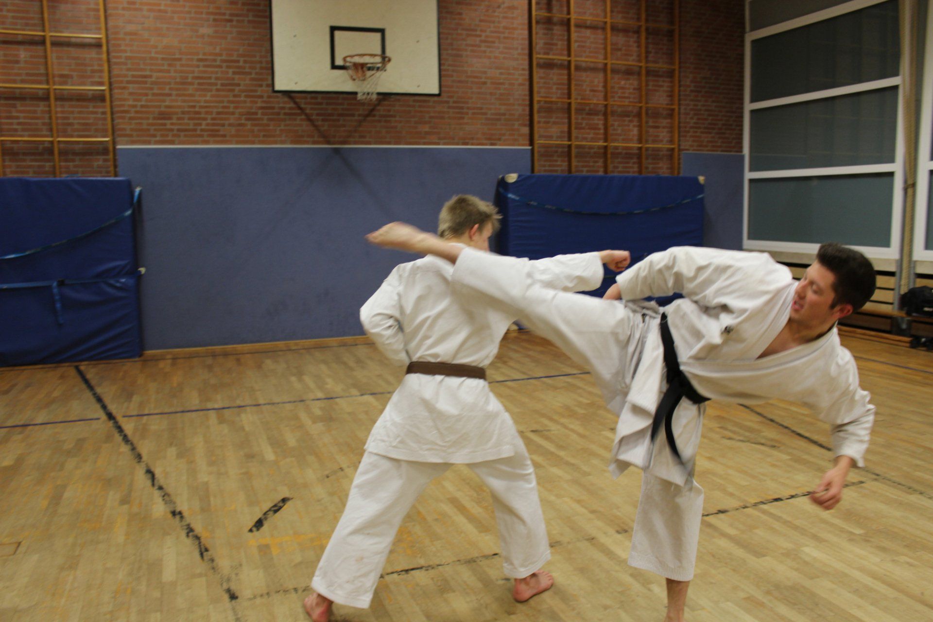 Zwei Männer üben Karate in einer Turnhalle . Im Hintergrund ist ein Basketballkorb zu sehen.