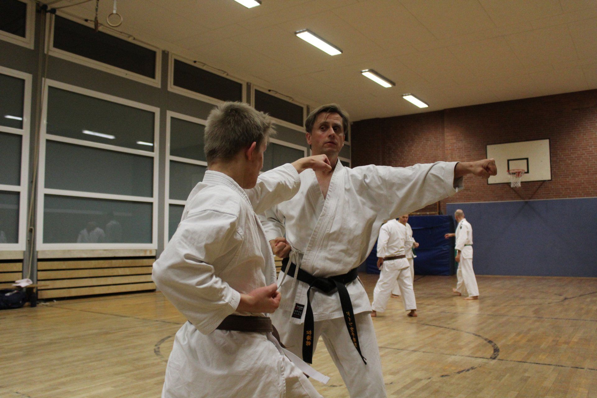 Zwei Männer üben Karate in einer Turnhalle mit einem Basketballkorb im Hintergrund