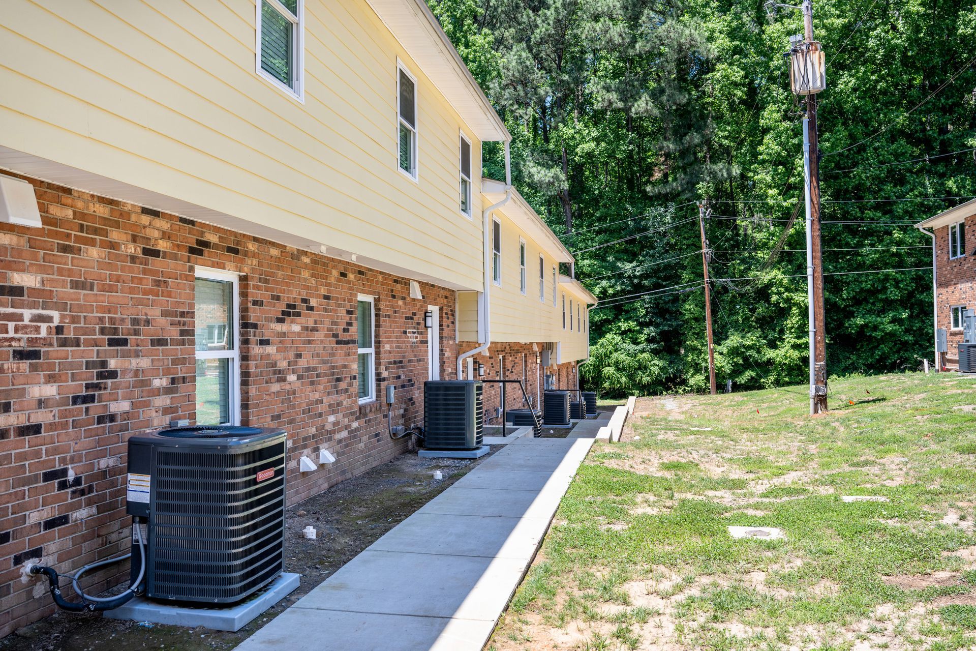 A sidewalk leading to a brick apartment building with air conditioners on the side of it.