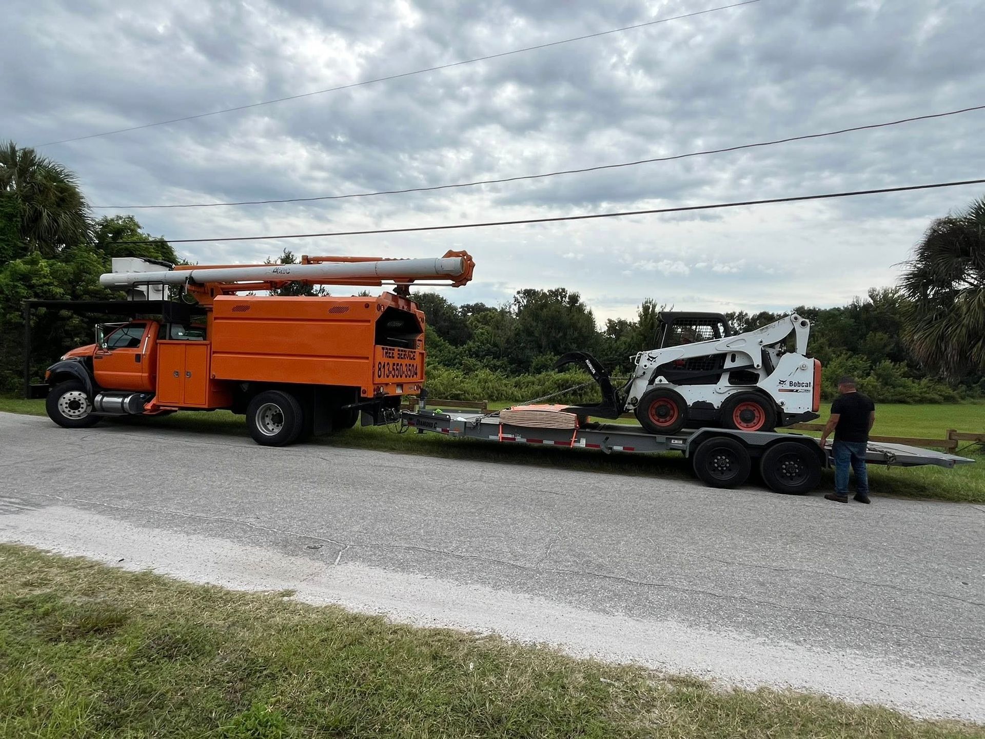 Orange truck towing a trailer with a Bobcat. A man stands near the vehicles on a road.