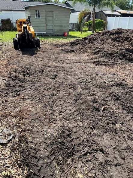 A large mulch pile and a stump grinder on a brown lot in front of a shed and fence.