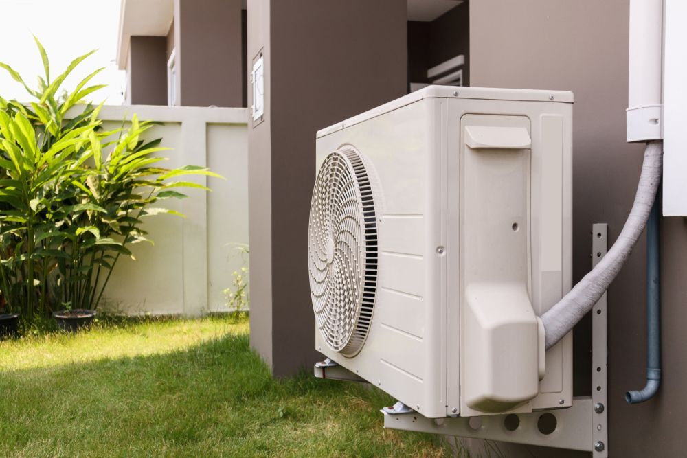 A Beige Air Conditioner Unit Mounted on a Grey Wall in a Backyard — Brian Williams Electrical Air Solar in Cooroibah, QLD