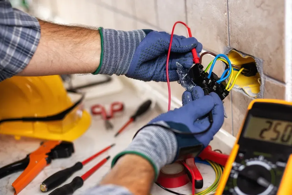 An electrician in blue gloves uses a multimeter to test electrical wires inside a wall outlet box. — Brian Williams Electrical Air Solar in Cooroibah, QLD