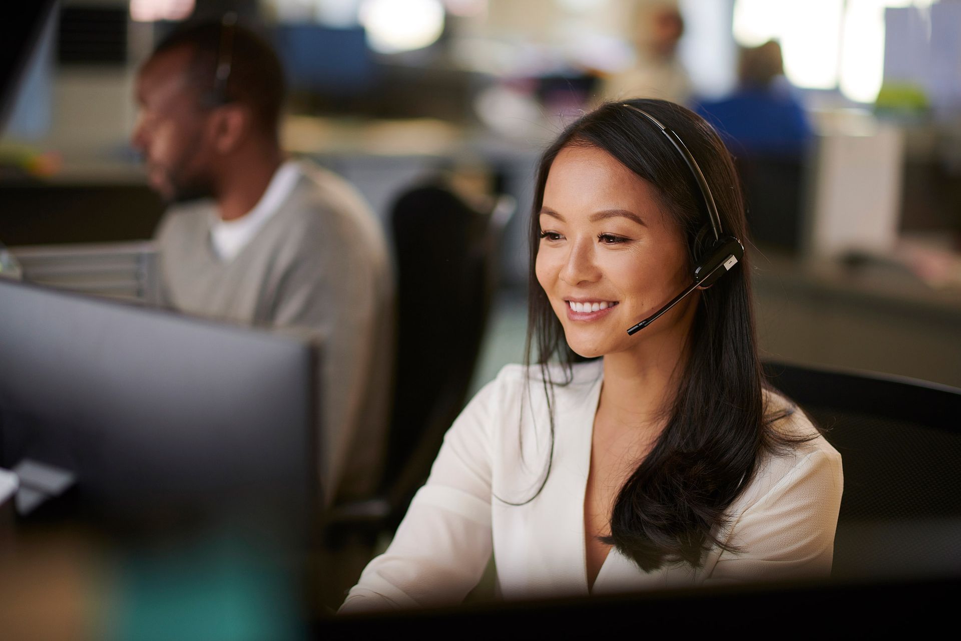 Woman with headset smiles while working on a computer in an office.