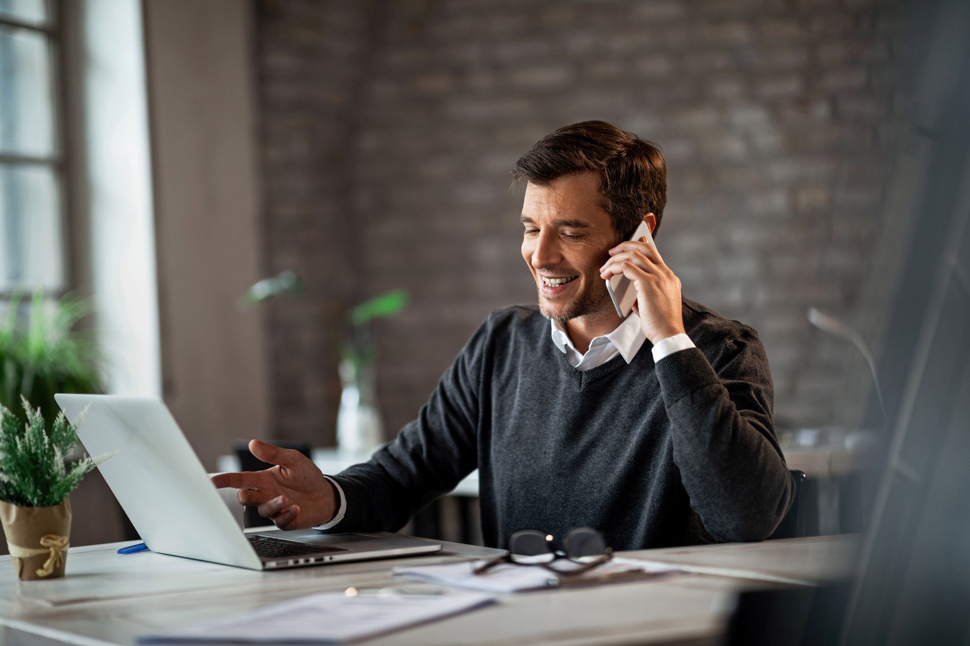 Man smiling while talking on the phone and working on a laptop at a desk.