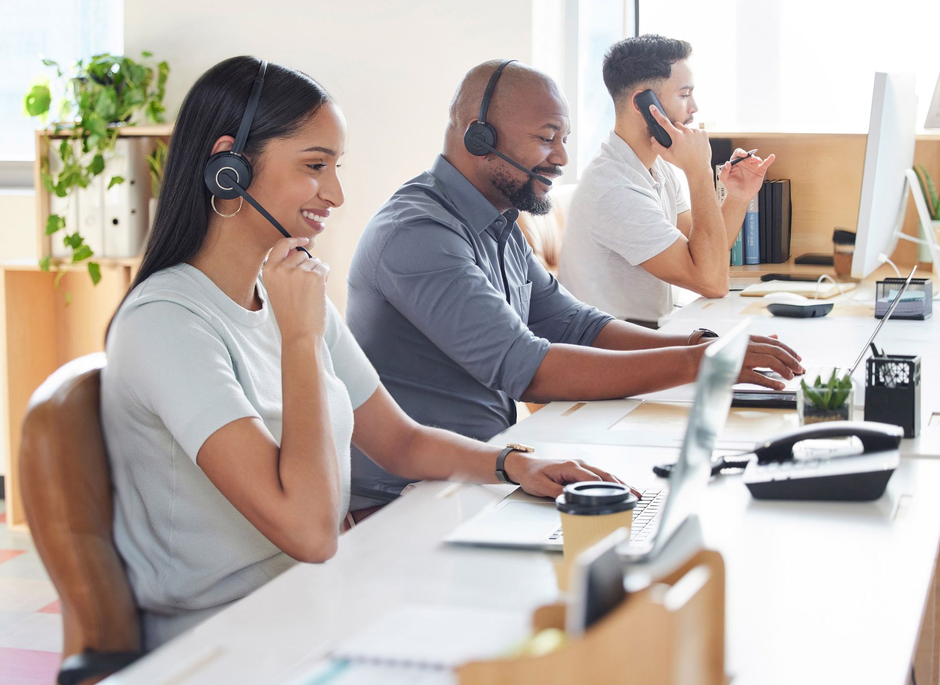 Three people wearing headsets working at desks with computers in an office setting.