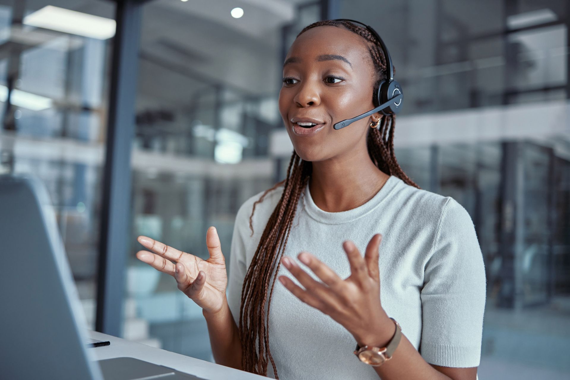Woman with headset talking on a video call, gesturing with hands, seated at a desk with a laptop, office setting.