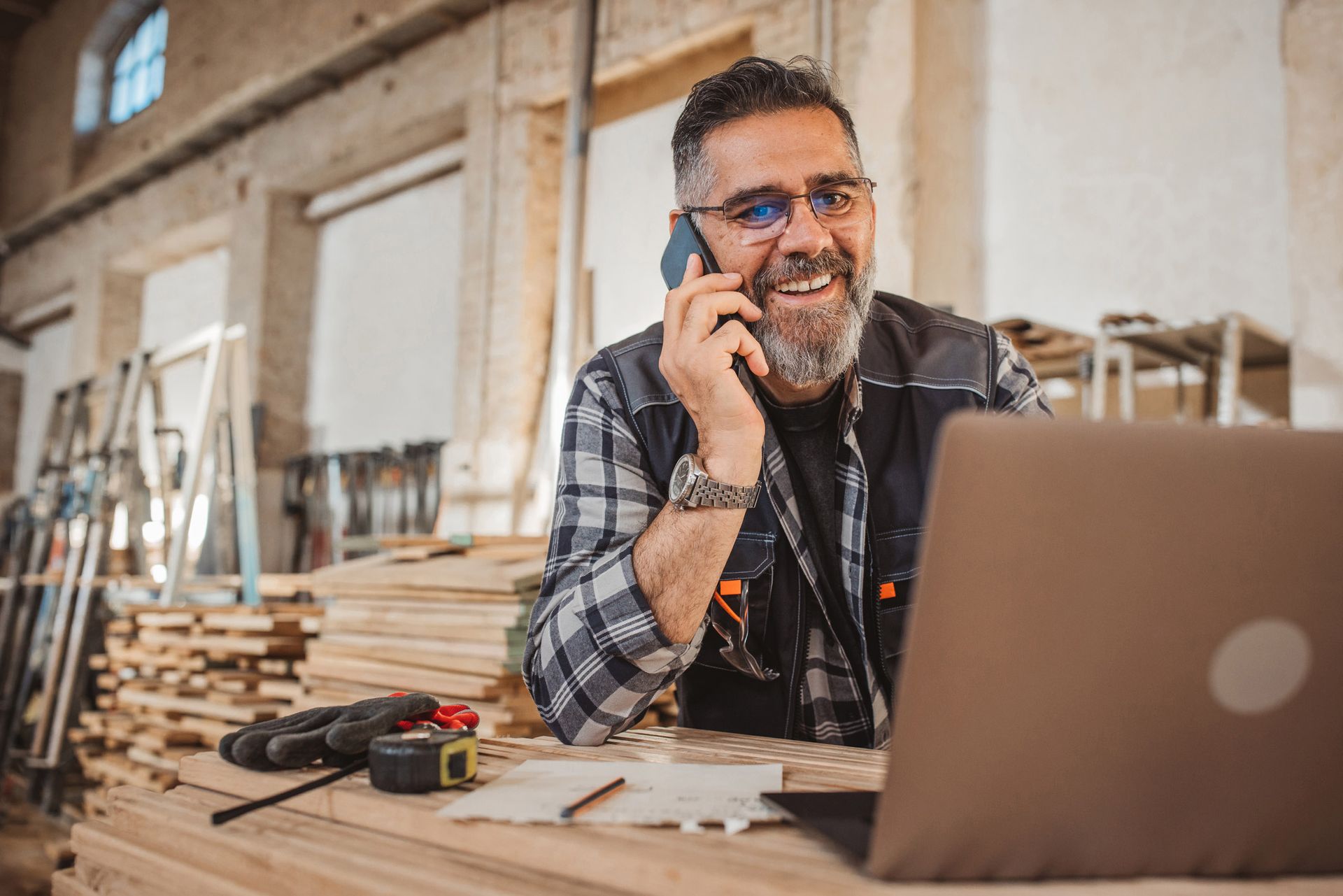 Man in workshop smiles while talking on phone, using laptop, surrounded by lumber and tools.