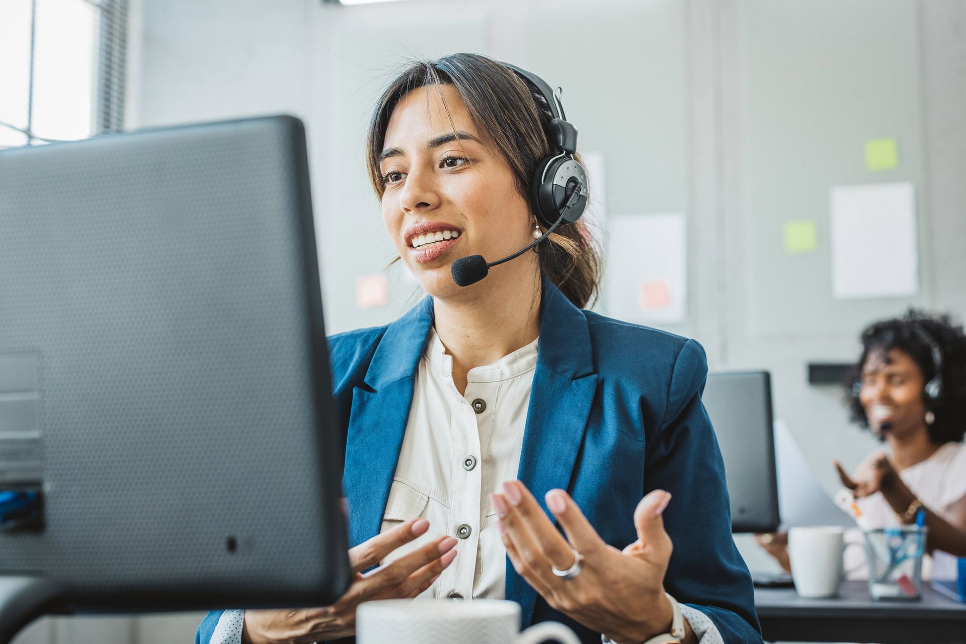 Woman in headset gestures at computer, smiling, in office setting.
