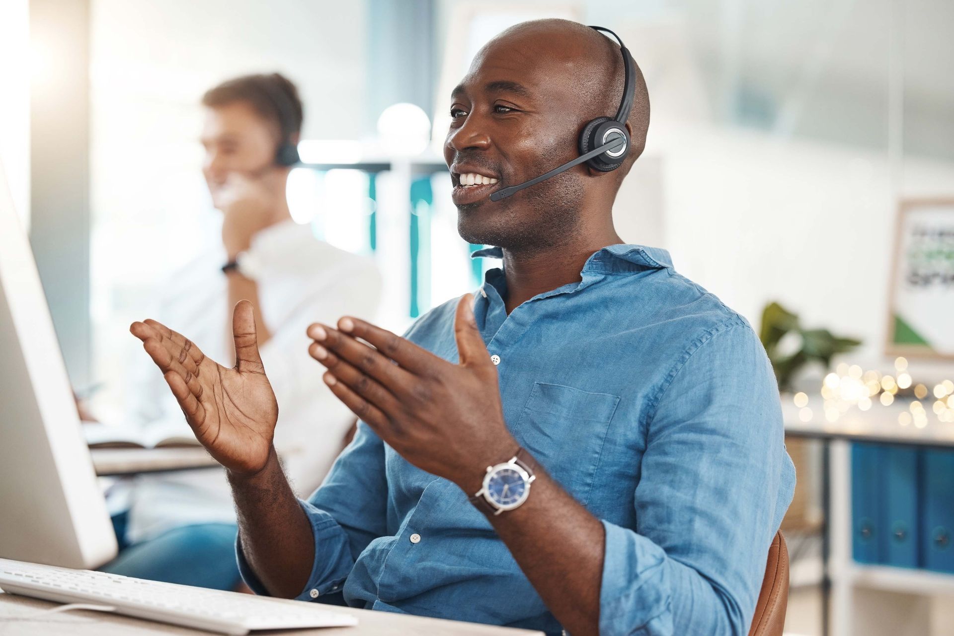 Man wearing a headset, gesturing with hands while talking at a computer in a brightly lit office.