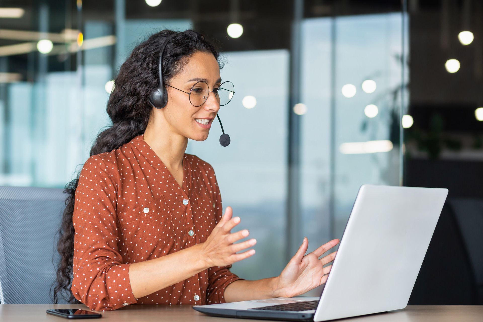 Woman with headset, smiling, using laptop in an office setting, gesturing with hands during a video call.