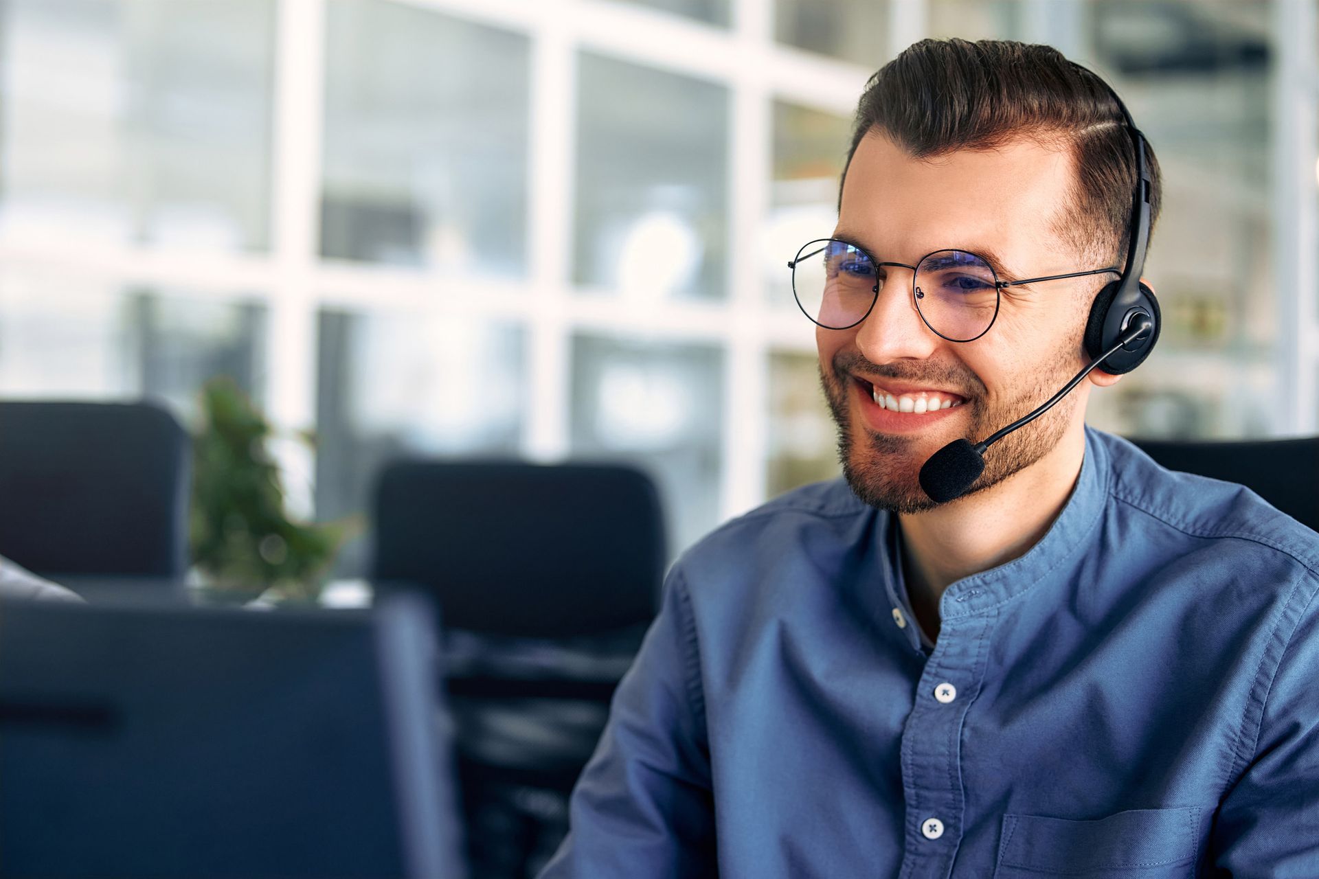 Man with headset and glasses, smiling while working at a computer in an office setting.