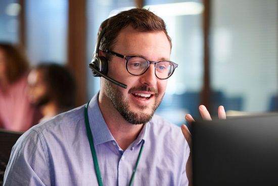 Man with glasses and headset smiles while looking at a computer screen in an office.