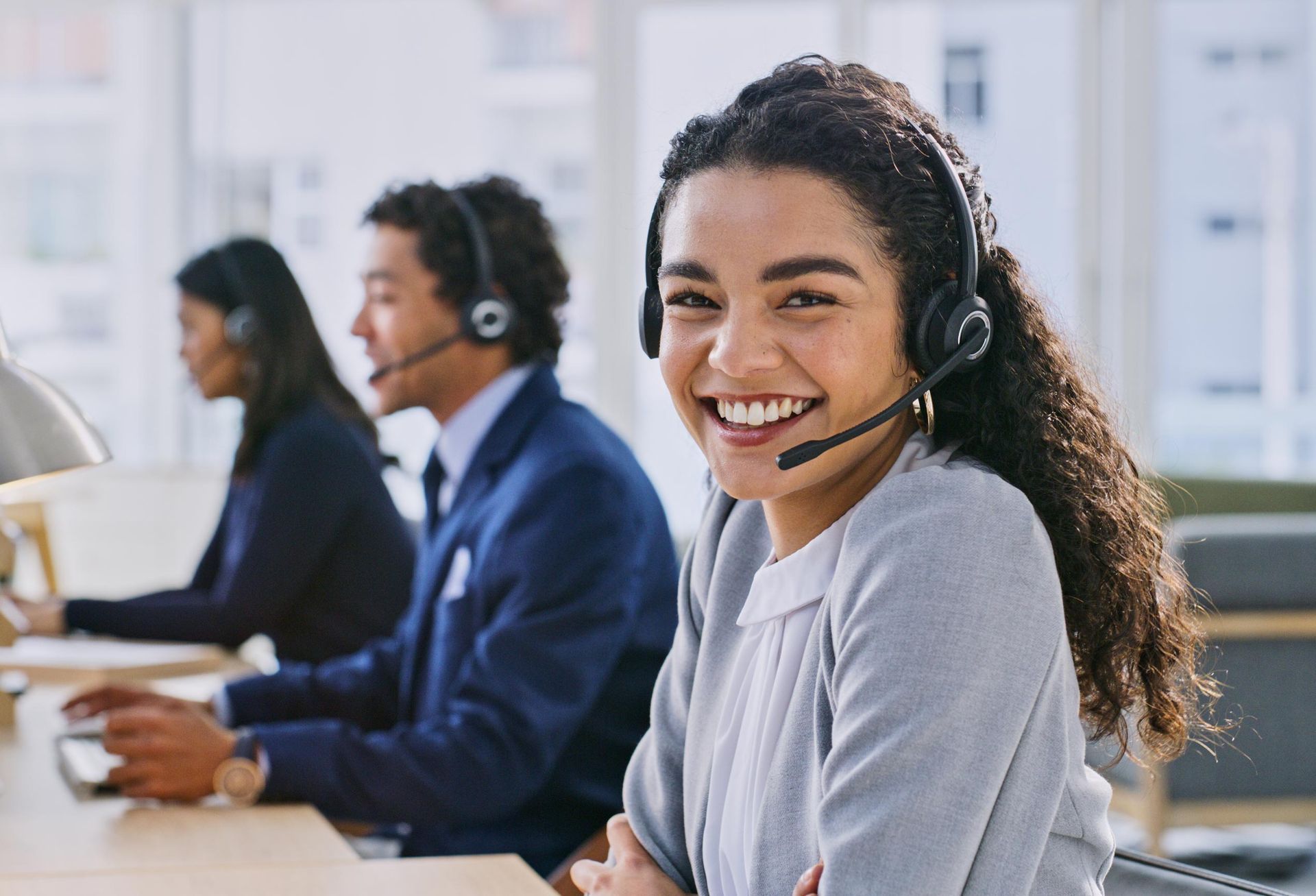 Woman wearing headset smiling at camera in office with coworkers.
