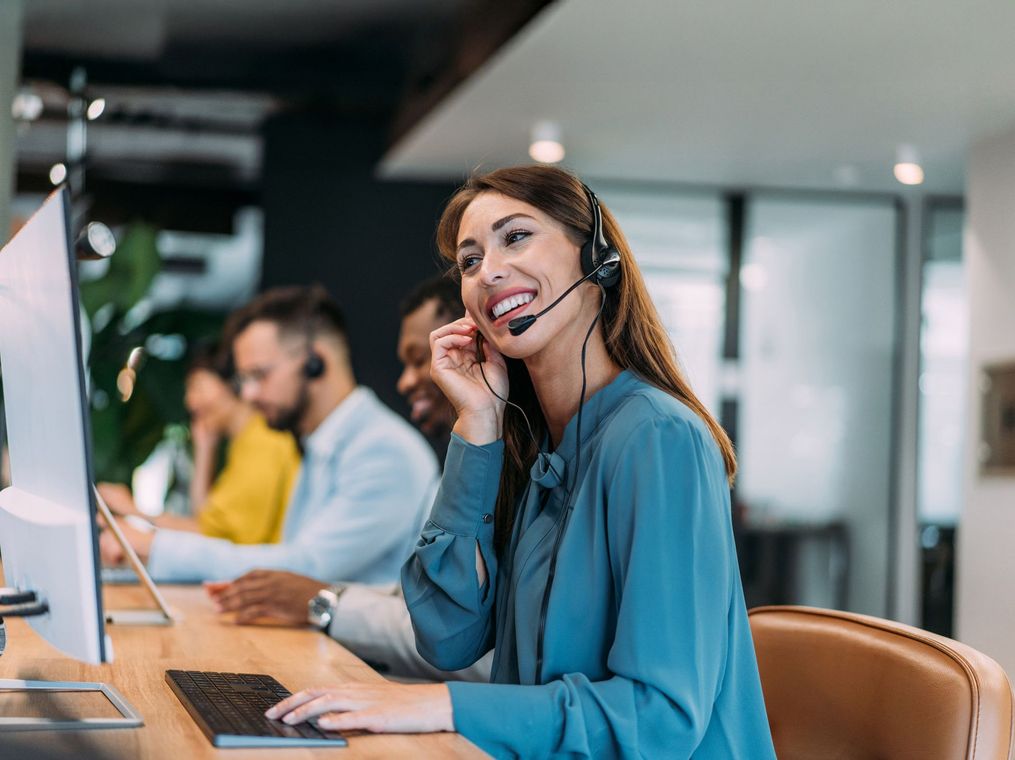 Woman with a headset smiling, working at a computer in an office setting. Other people are in the background.