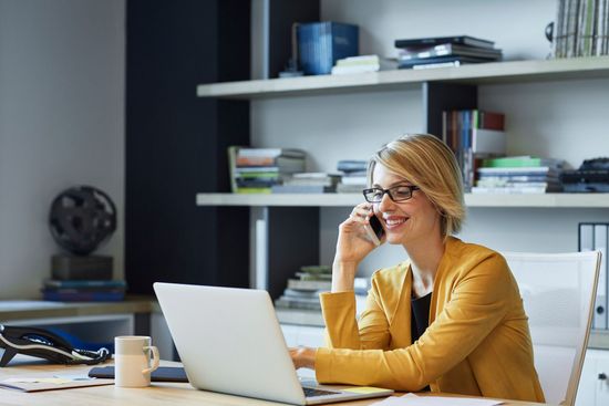 Woman in yellow blazer on phone, using laptop at desk, bookshelves in background.