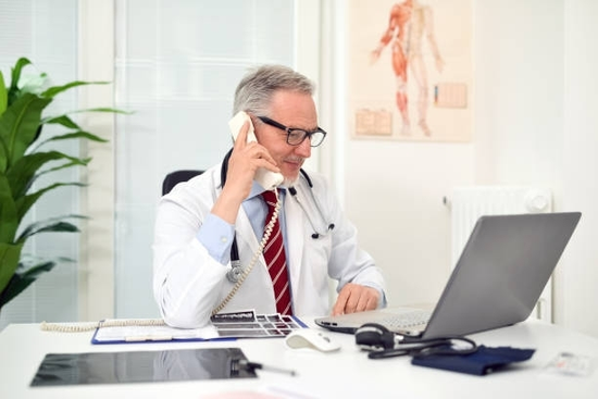 Doctor on a landline phone, looking at a laptop, stethoscope around his neck, in a bright office.