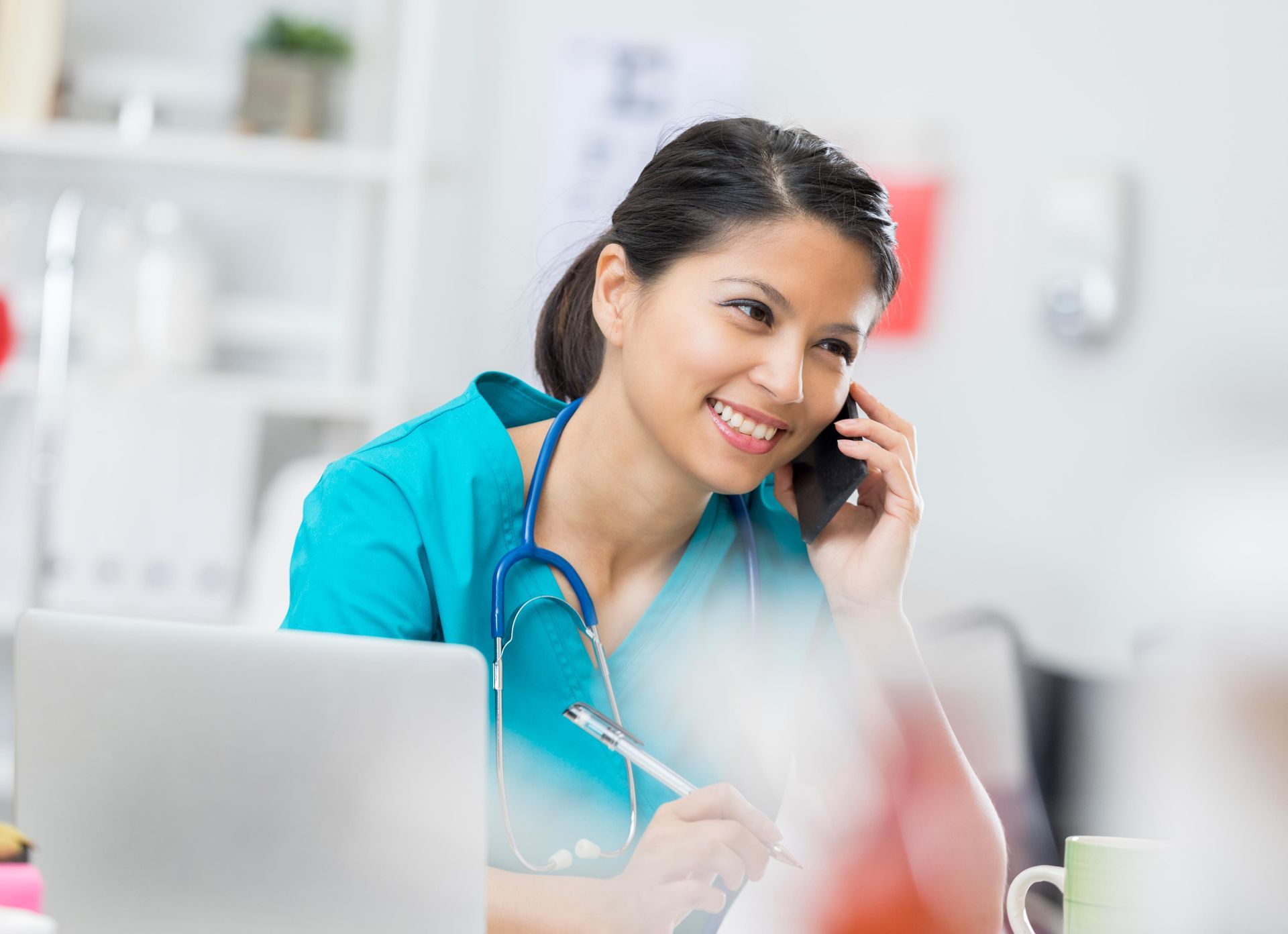 Nurse in teal scrubs smiles while talking on phone in a medical office with laptop and stethoscope.