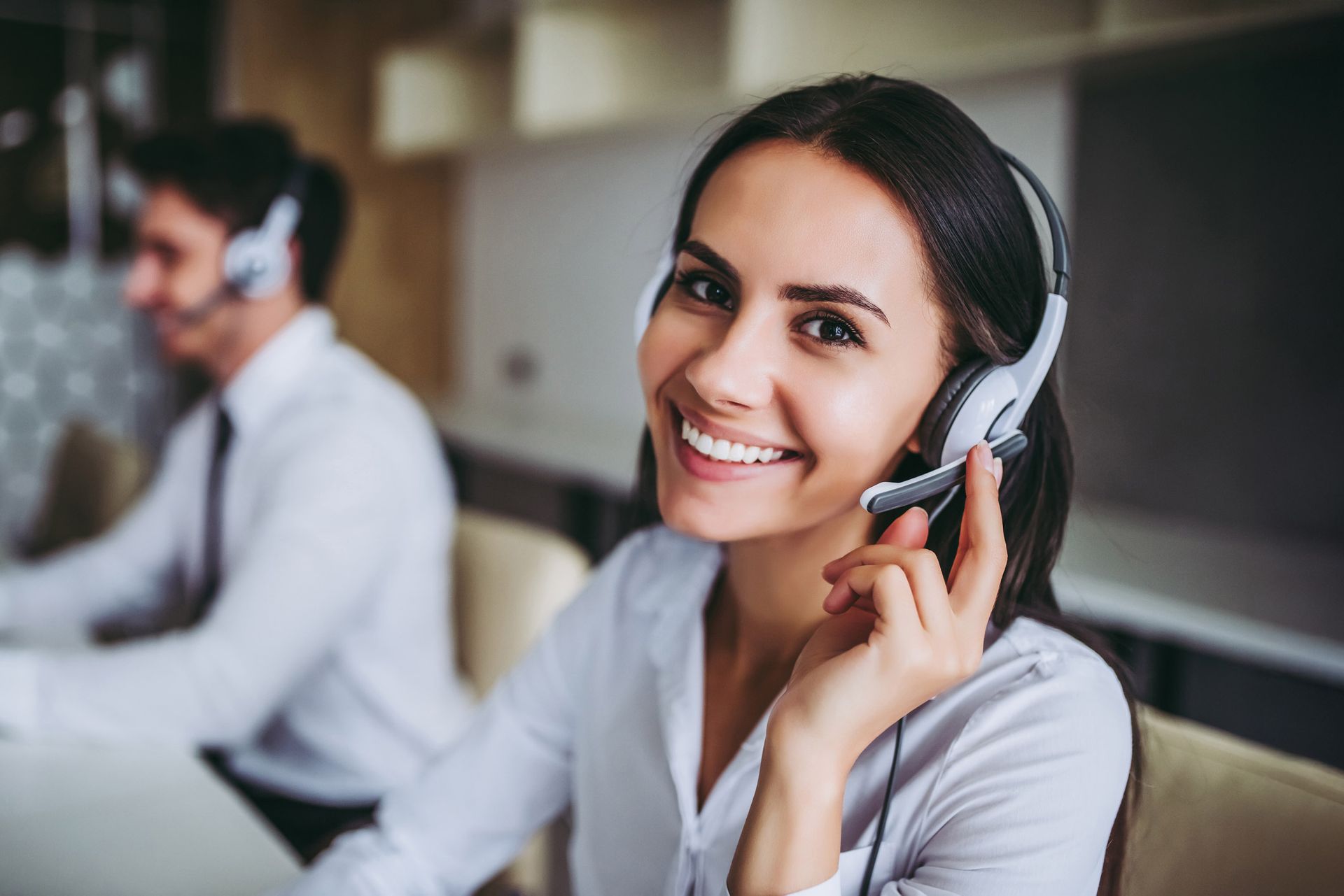 Woman with a headset smiles, in an office setting. A man in the background also wears a headset.