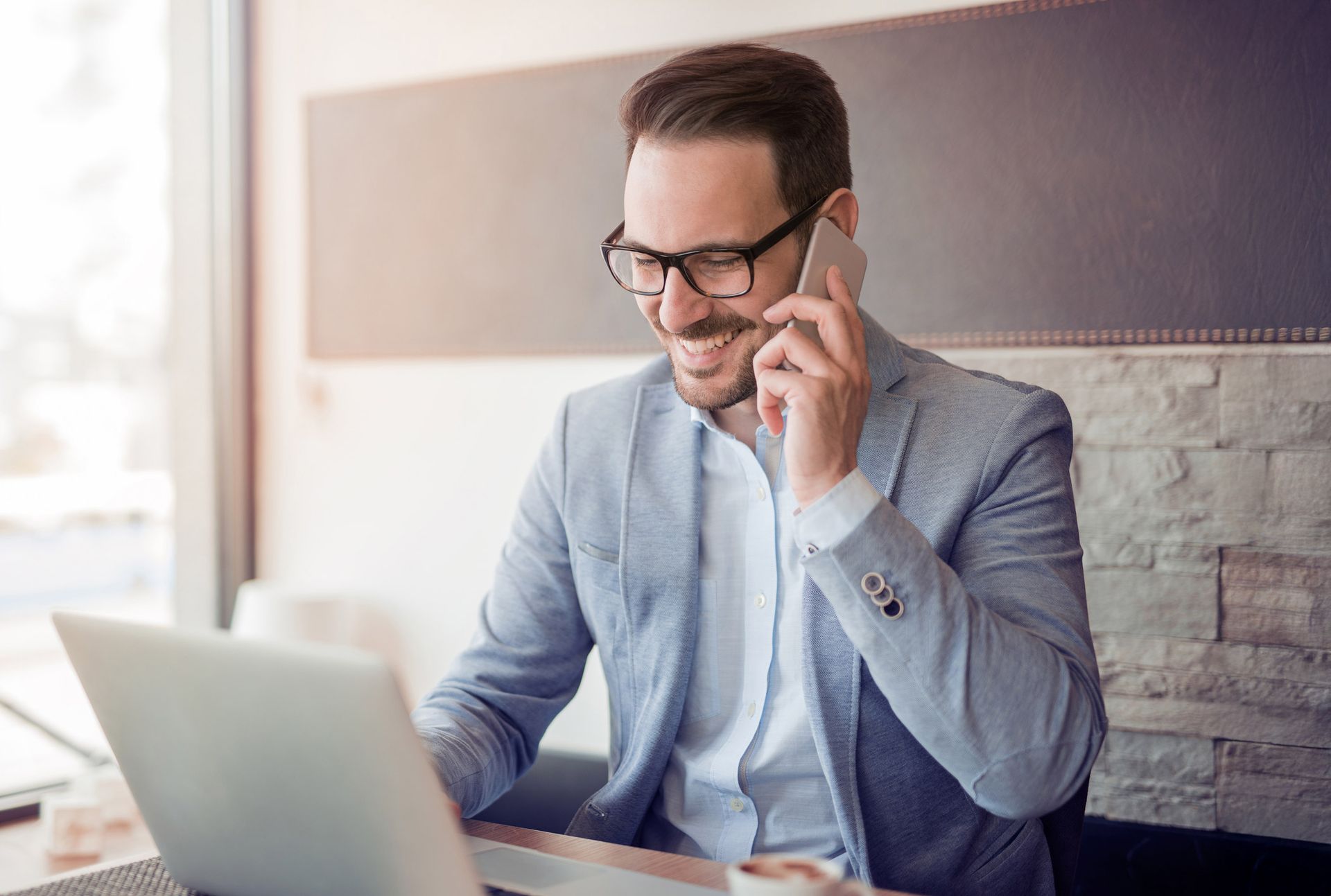 Man in blazer smiling, on phone, working on laptop in cafe.