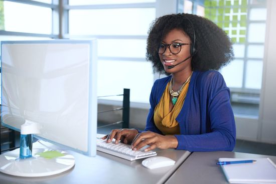 Woman with headset typing at computer in an office setting, smiling.