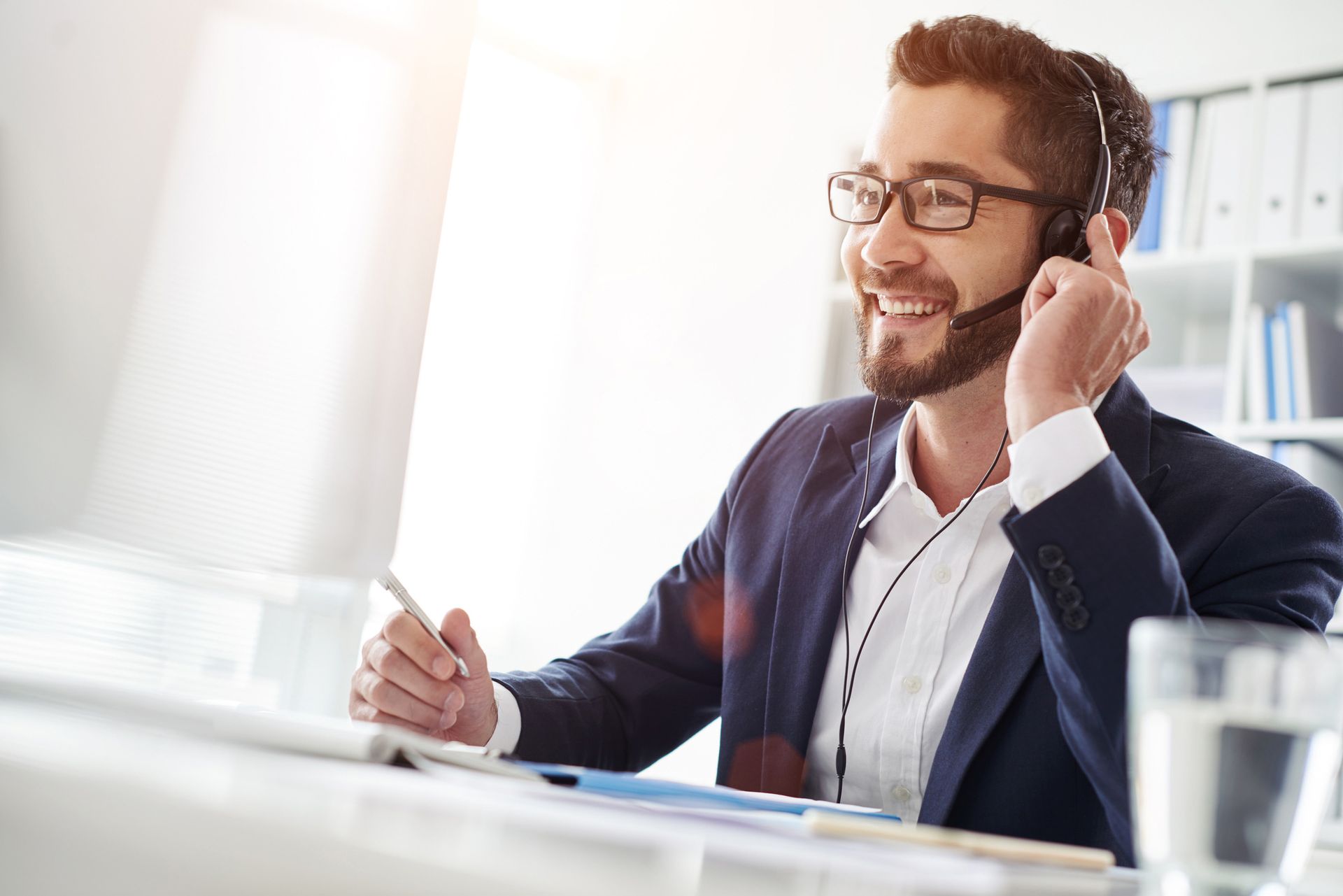 Man in blazer with headset smiles while working on a computer at a desk.