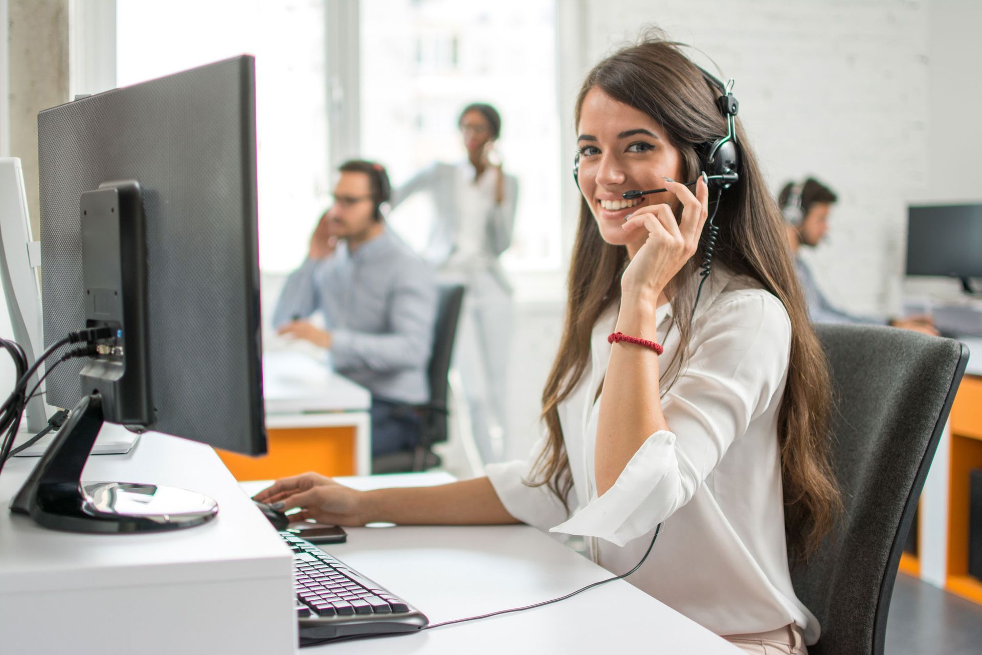 Woman with headset smiles at the camera while working at a computer in an office setting.
