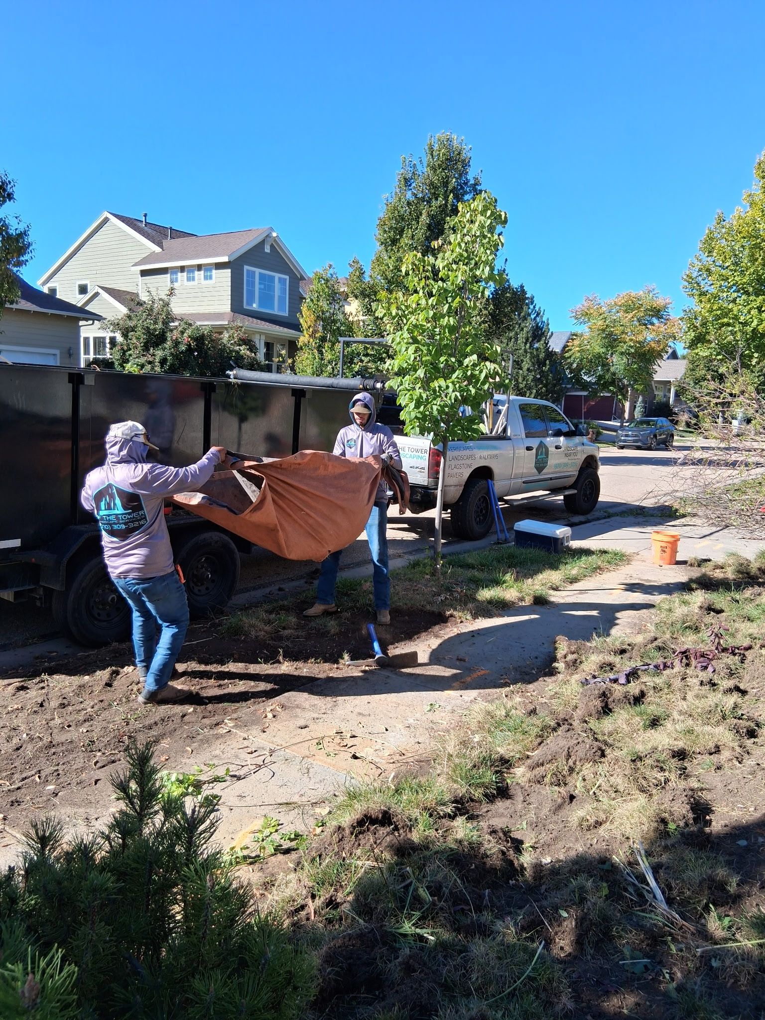 Two people emptying debris from a truck on a sunny day. Houses and a white truck are in the background.