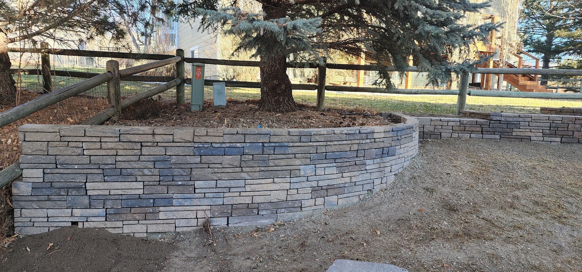 A stone retaining wall curves around a tree base in a yard, with a fence in the background.