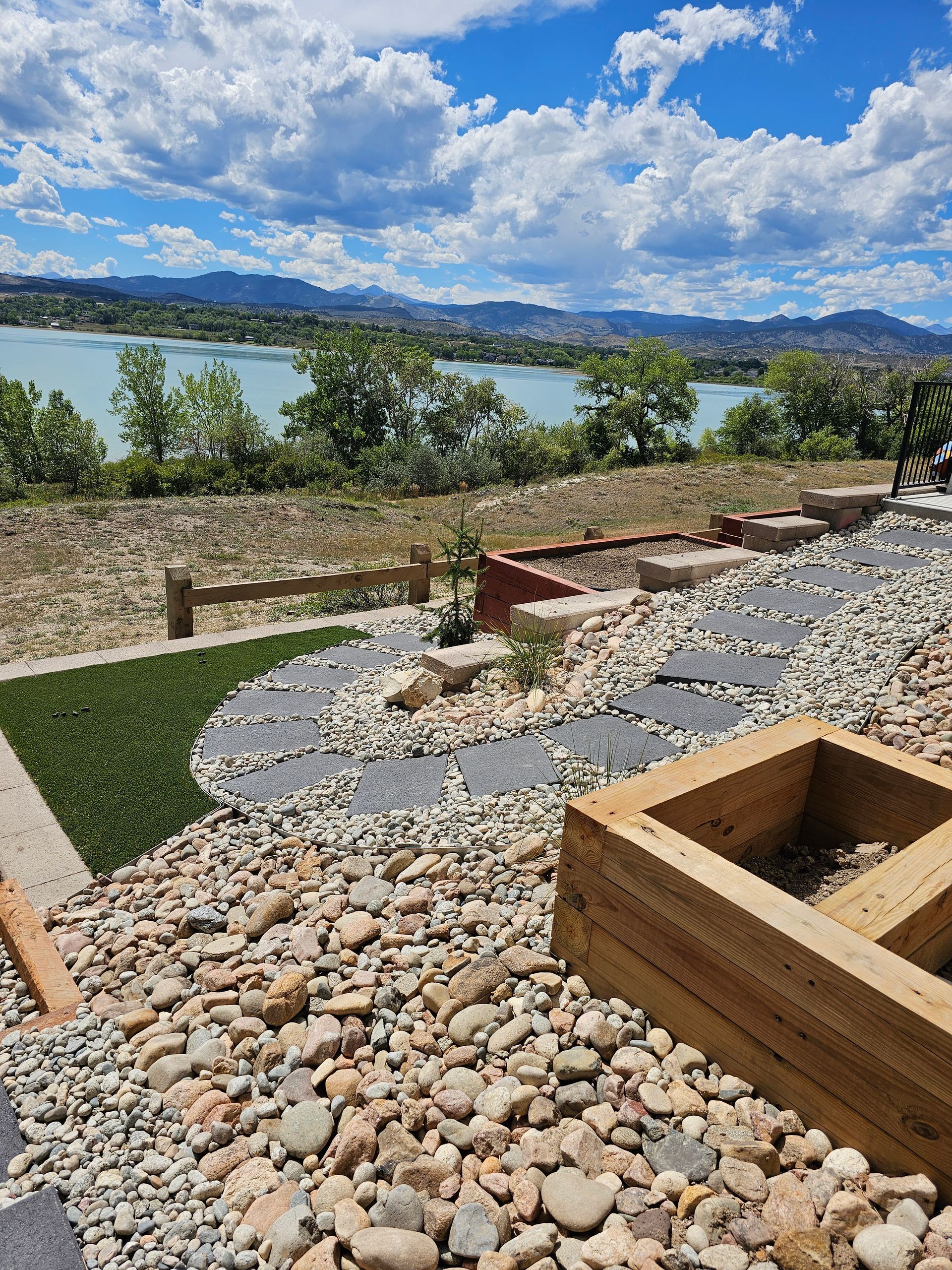 A scenic outdoor patio with river view; gray stone path, wooden planter boxes, blue sky with fluffy clouds.