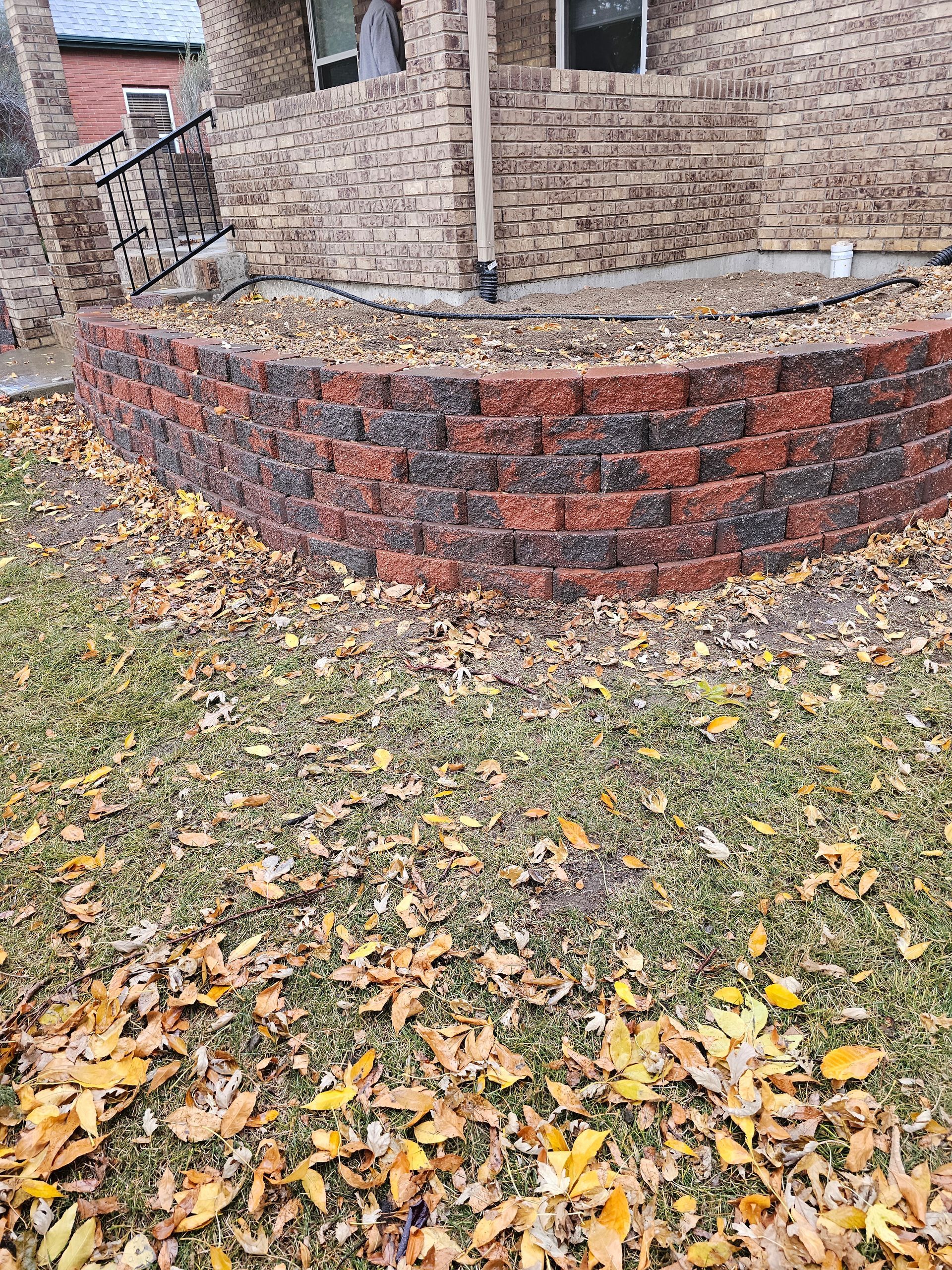 Brick retaining wall with brown and red bricks, fronting a house with fallen leaves on the grass.