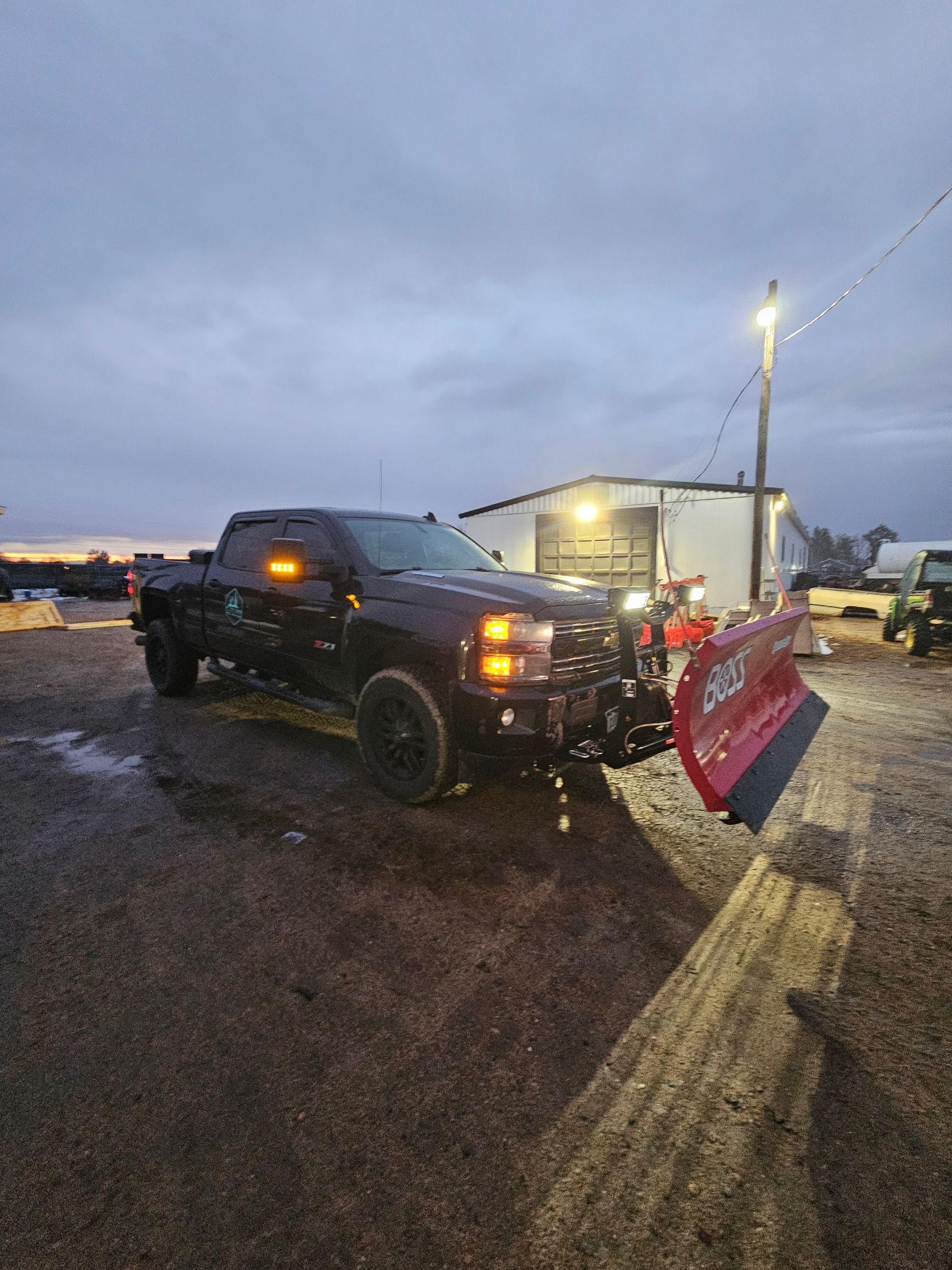 Black pickup truck with snowplow, parked on gravel, cloudy sky.
